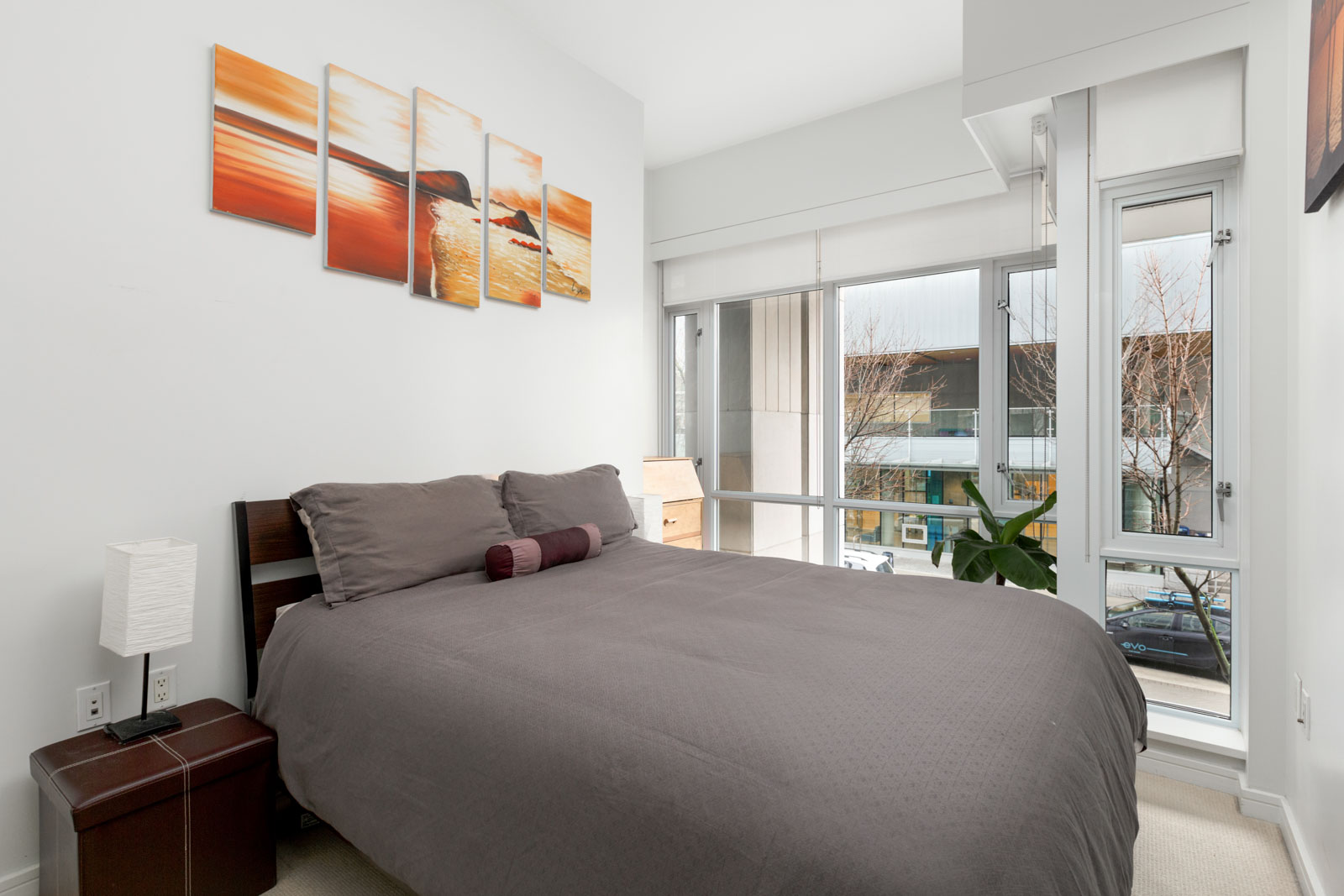 queen size bed with dark grey sheets in condo bedroom with painting above and floor to ceiling windows on right looking outside