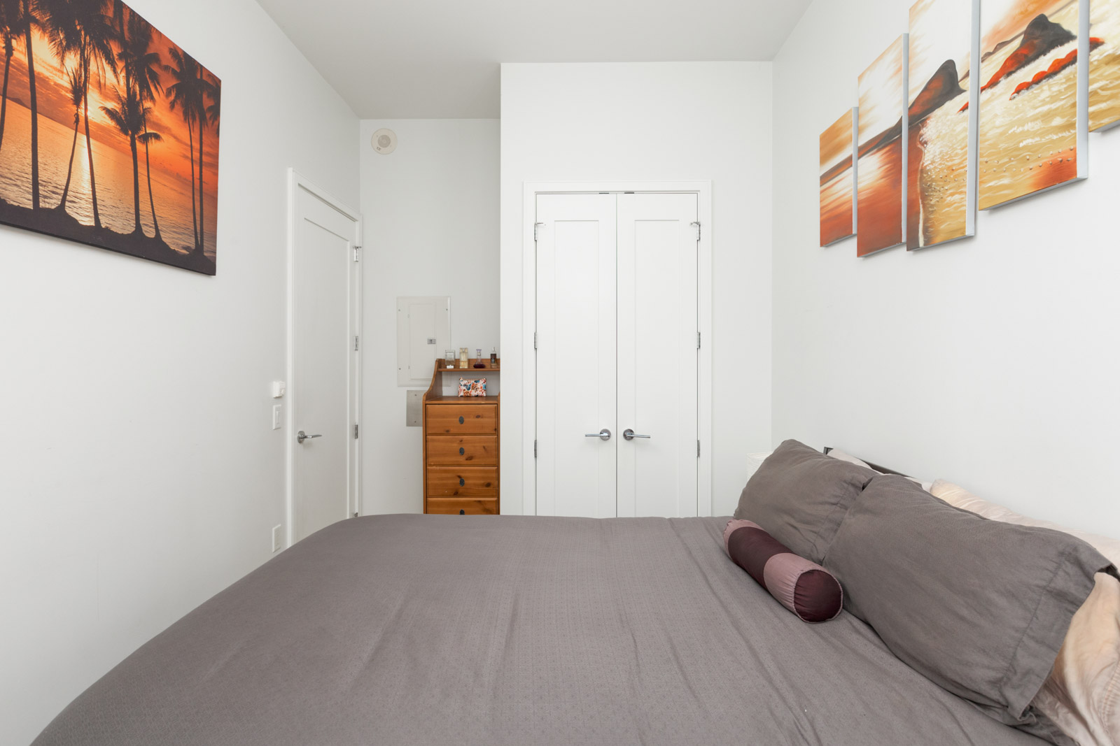 condo bedroom with bed with grey sheets in foreground and closed door closet space in background with white doors and white walls