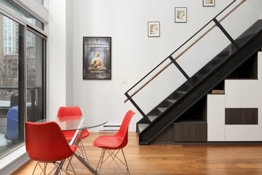 side view of dark stairs with railing to upstairs of loft apartment with red dining chairs on left and white wall in the back