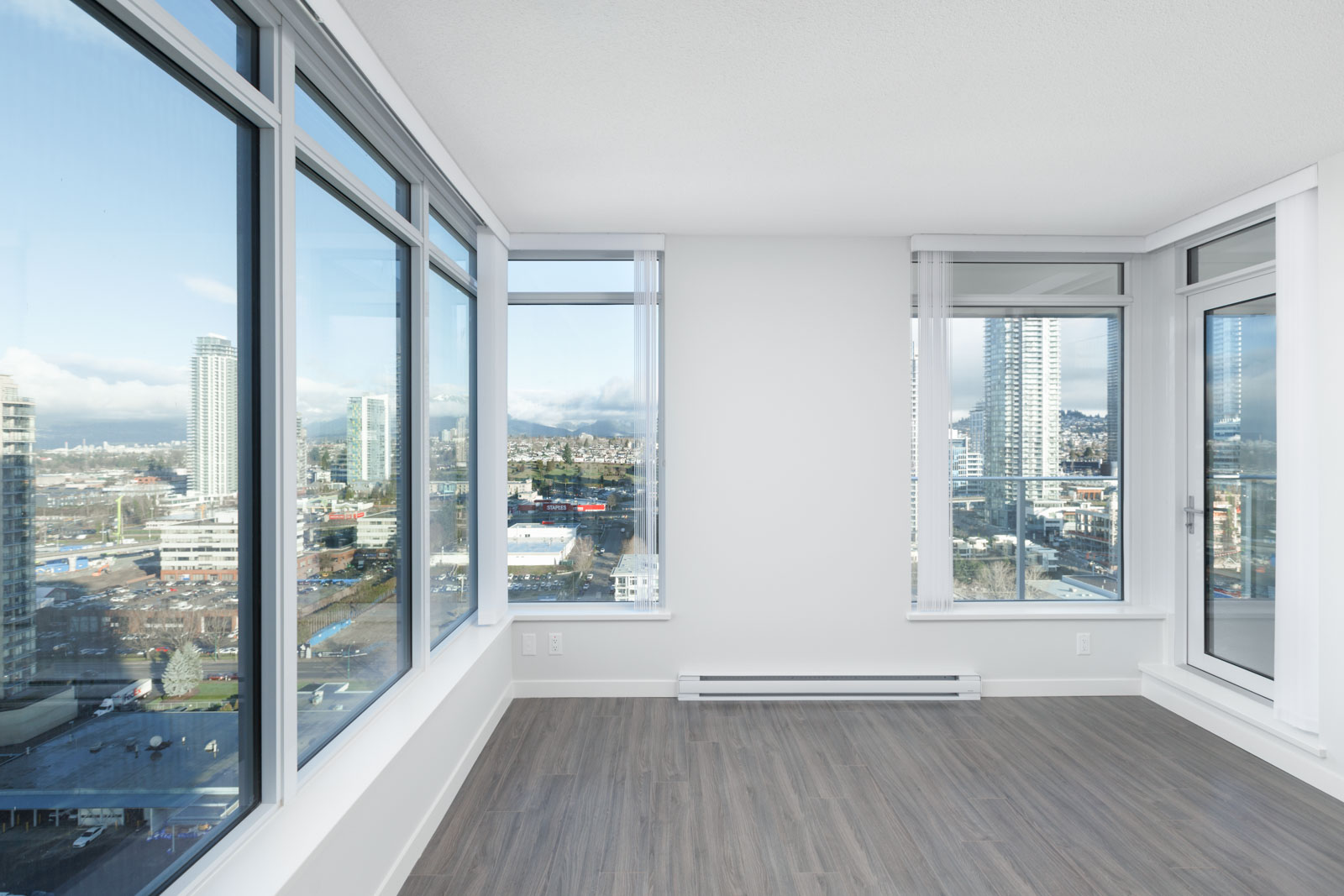 Empty modern apartment room with large floor-to-ceiling windows, light-colored walls, and wood flooring, overlooking a cityscape with tall buildings and blue sky.