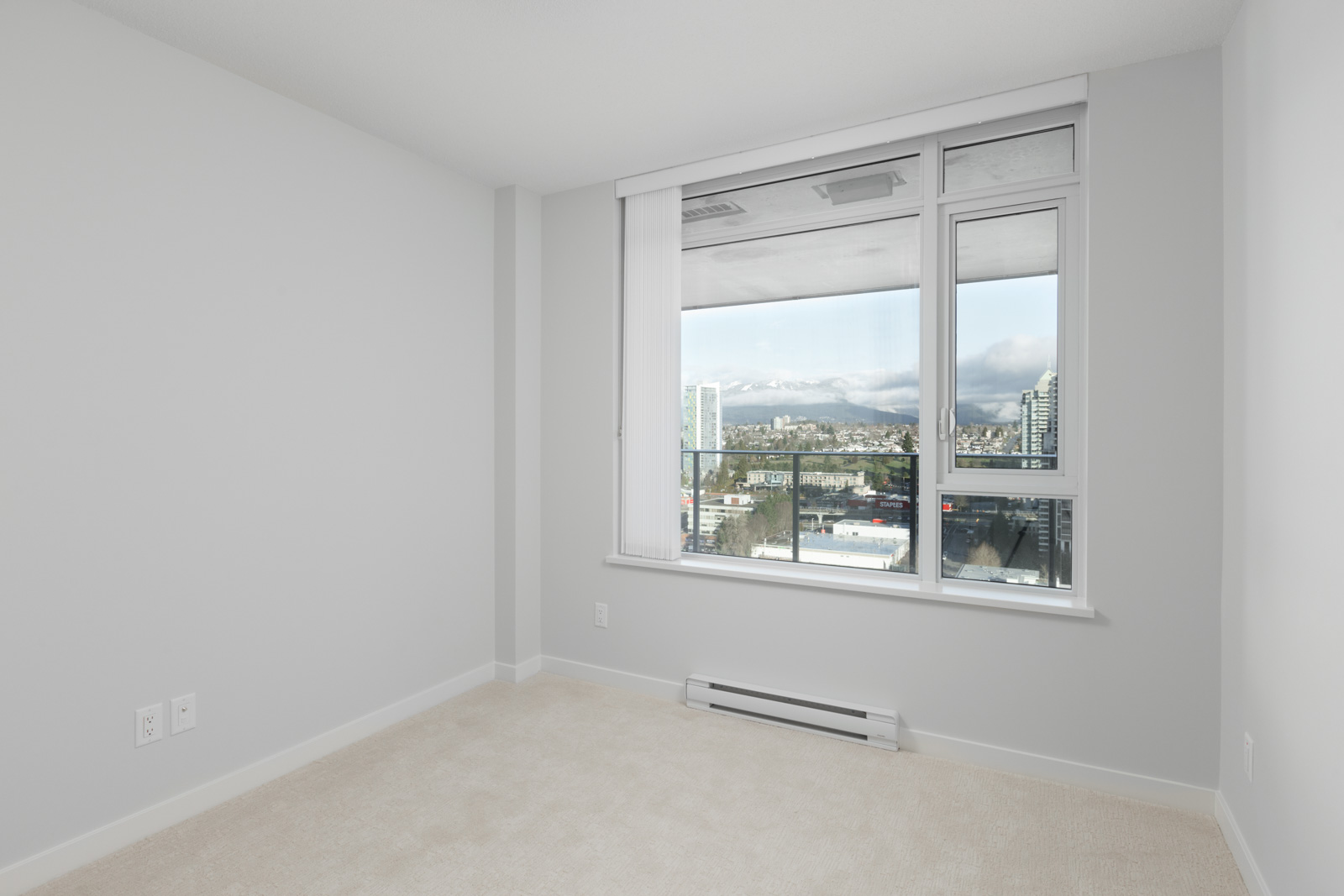 Empty room with light-colored carpet, white walls, and a large window showing a cityscape and mountains in the background.