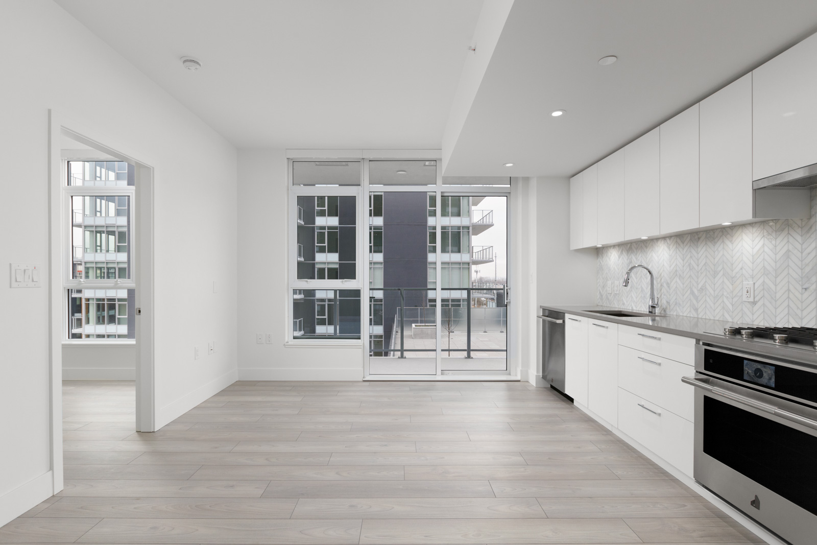 Modern, unfurnished apartment kitchen with white cabinets, stainless steel appliances, light wood flooring, and large windows overlooking a balcony and neighboring building.