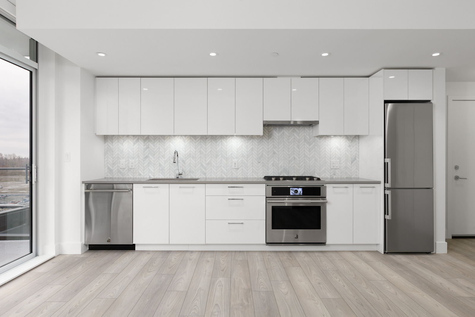 Modern kitchen with white cabinets, stainless steel appliances, light wood floor, and a herringbone tile backsplash. Large window on the left provides natural light.