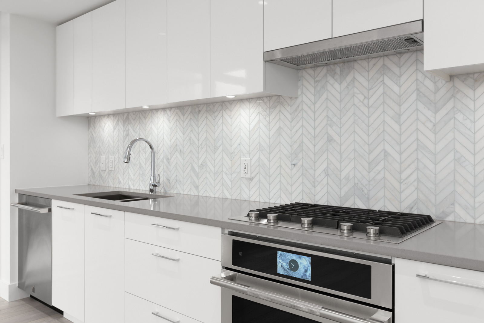Modern kitchen with white cabinets, gray countertop, stainless steel gas stove and oven, built-in dishwasher, sink with faucet, and light herringbone tile backsplash.