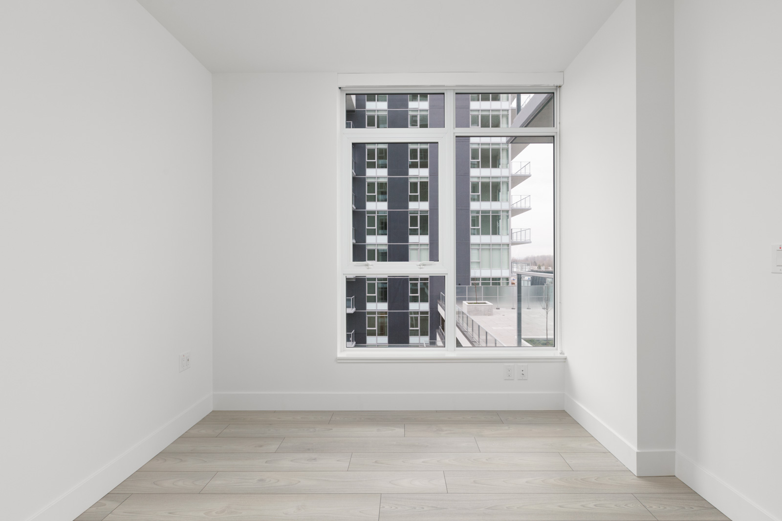 Empty white room with light wood flooring, a large window, and a view of modern apartment buildings outside.