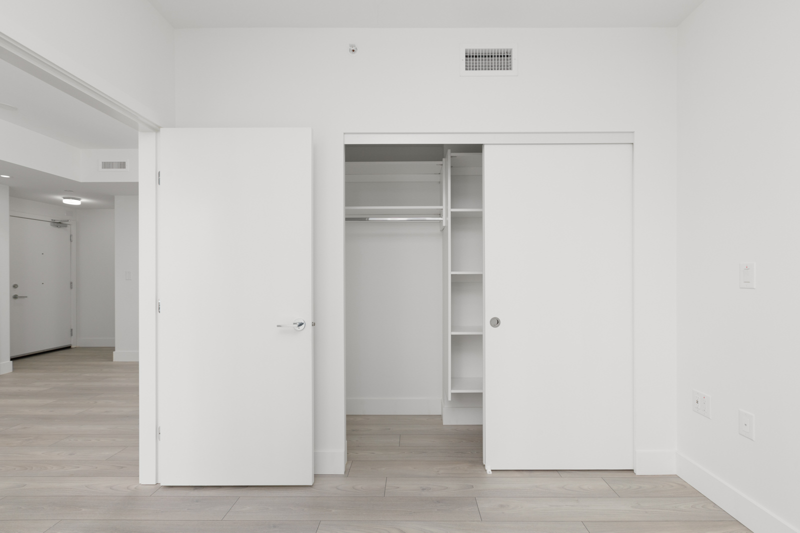 Open closet with sliding doors in a white, empty room with light wood flooring and built-in shelves. Adjacent door leads to hallway with visible front door.