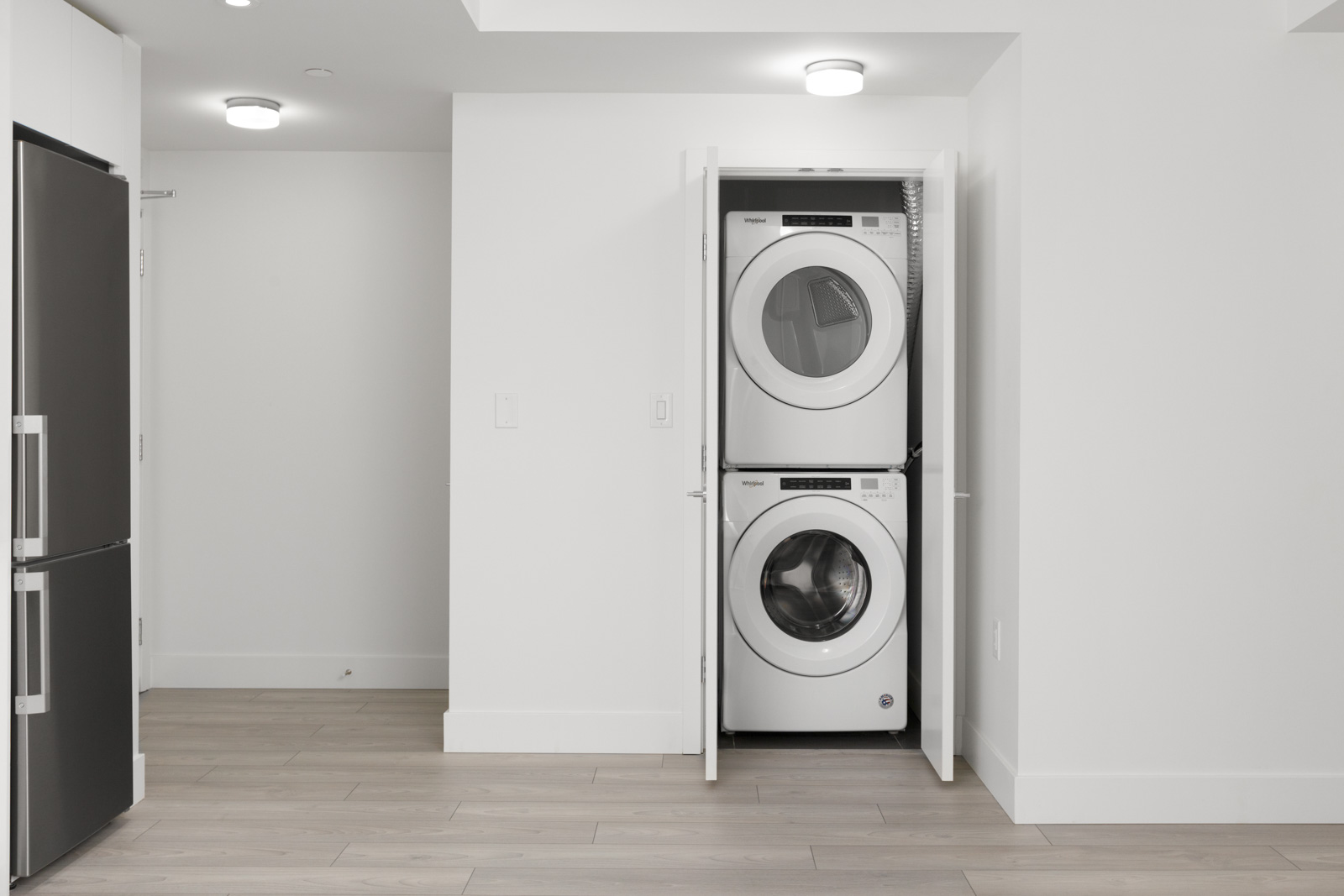 A stacked washer and dryer set is enclosed in a white closet in a modern, minimalistic laundry area with light wood flooring.