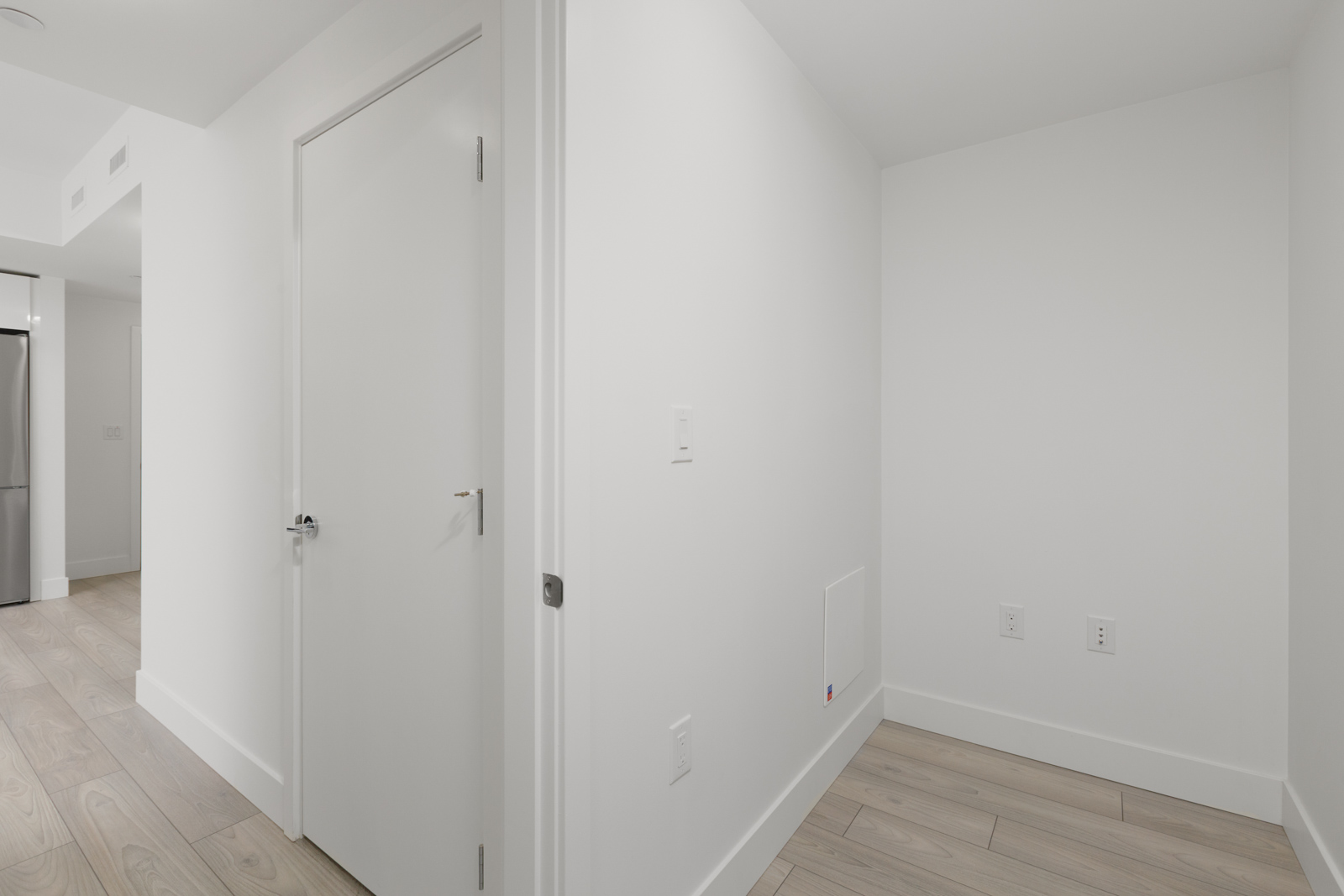 A small, empty white room with light wood flooring, white walls, a closed door, and electrical outlets on the right wall. A kitchen area is partially visible in the background.