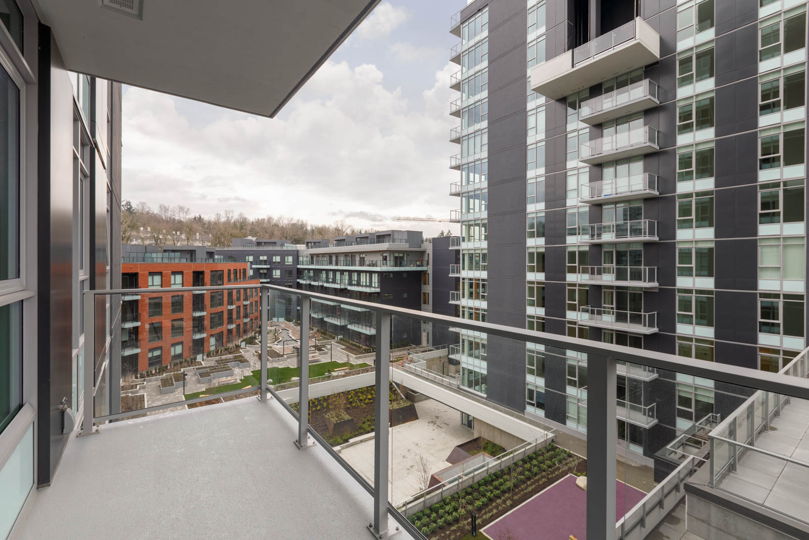 View from a balcony overlooking a modern apartment complex with multiple buildings, glass railings, landscaped courtyard, and cloudy sky.