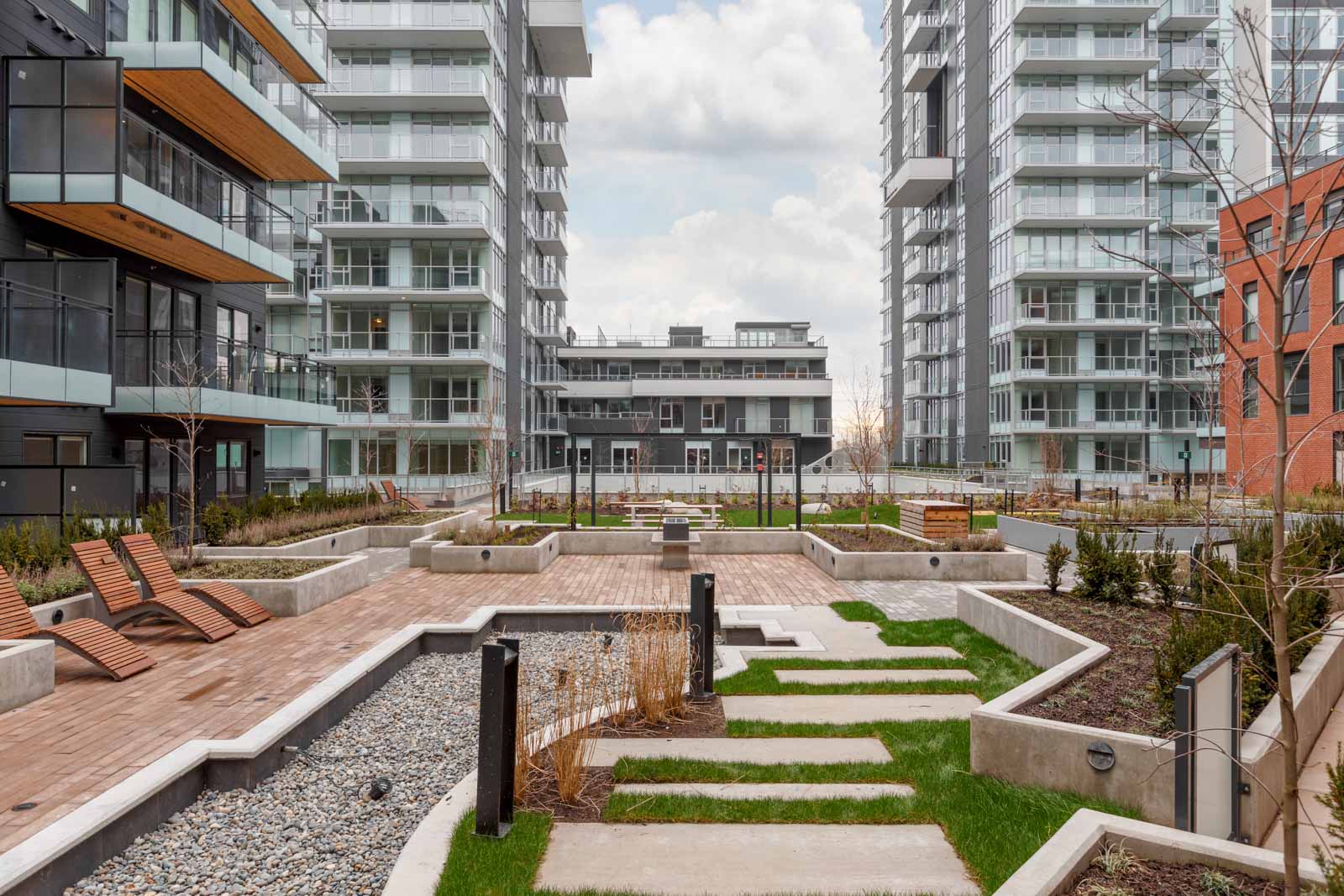 Modern apartment complex courtyard with lounge chairs, paved walkways, landscaped garden beds, and surrounding high-rise buildings under a cloudy sky.