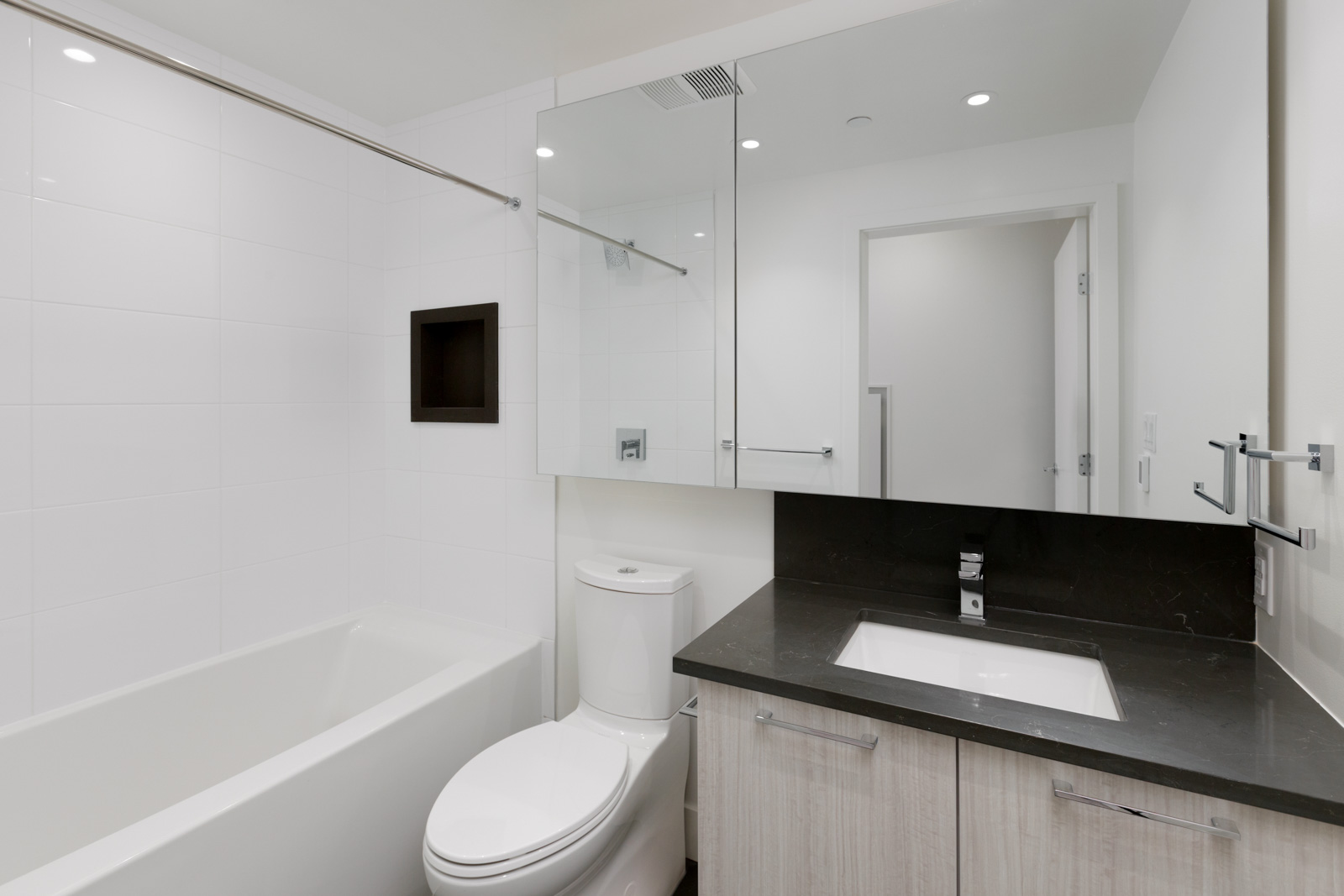 Modern bathroom with a bathtub, toilet, and sink; features a large mirror, white walls, black countertop, and light wood cabinetry.