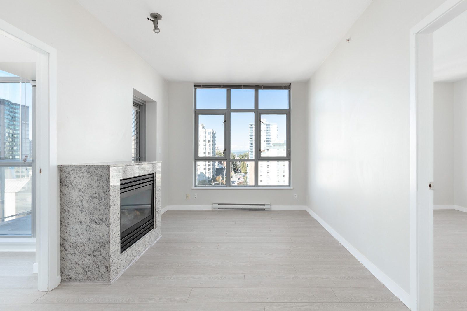 Bright, unfurnished apartment living room with large windows, a modern fireplace, light wood flooring, and white walls. Two open doorways lead to adjacent rooms.