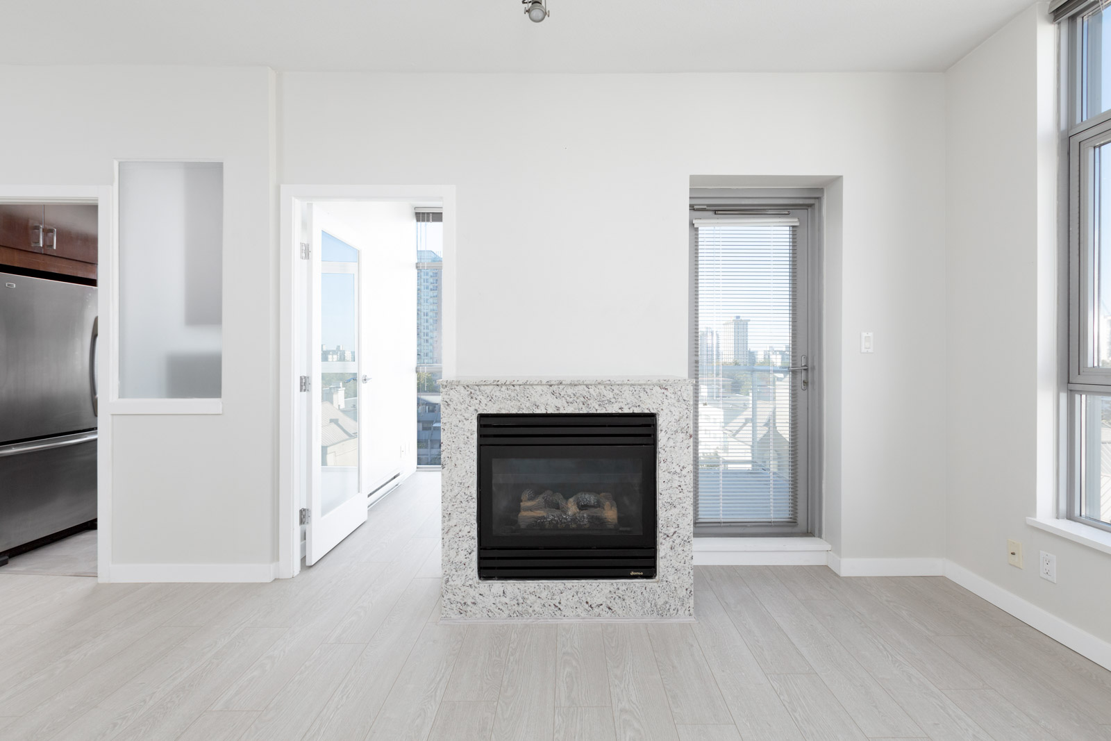 Modern living room with light wood flooring, a central fireplace, large windows, a glass door to a balcony, and partial view of a kitchen with stainless steel appliances.
