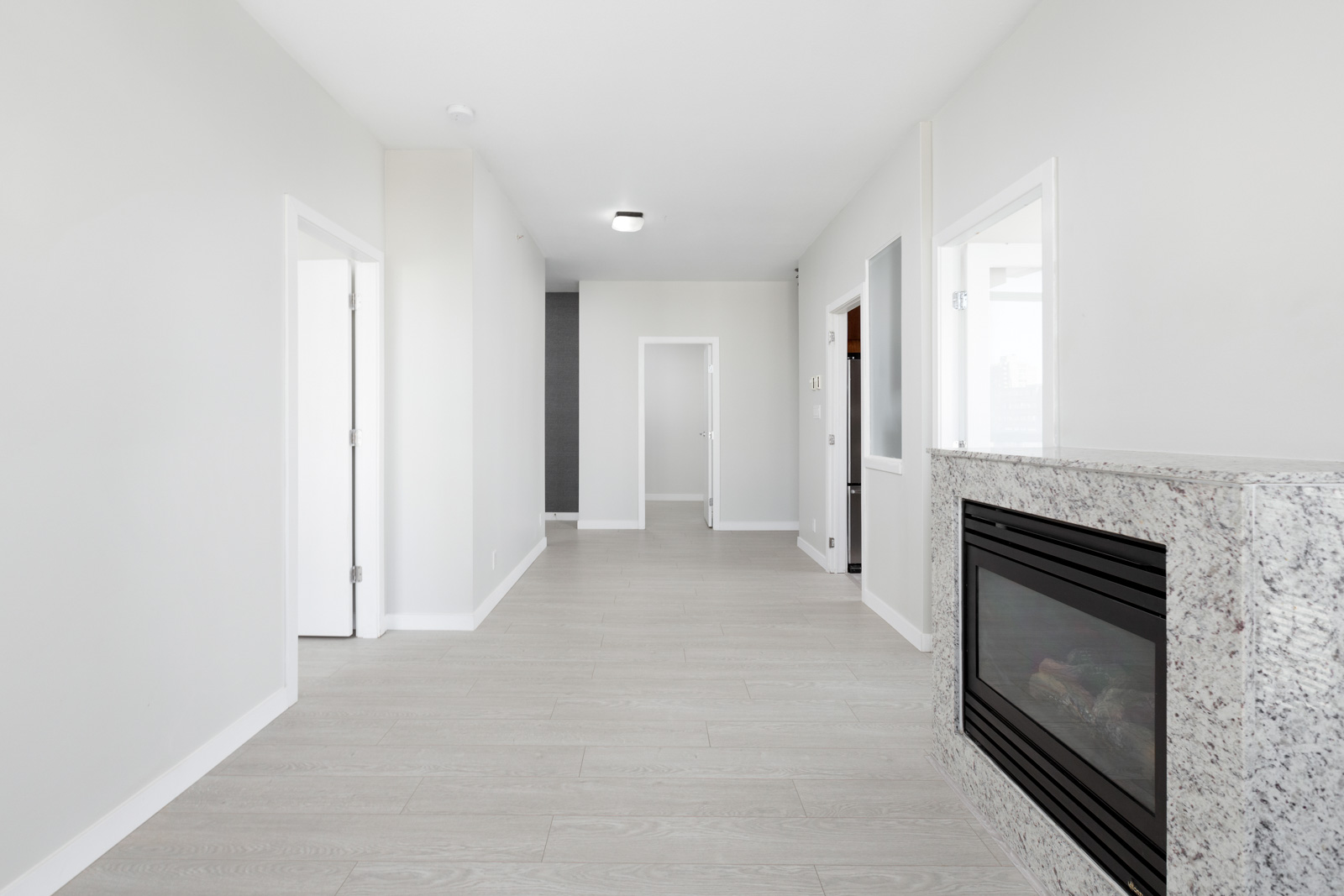 A modern, unfurnished room with light wood flooring, white walls, a granite fireplace on the right, and several doorways leading to other rooms.
