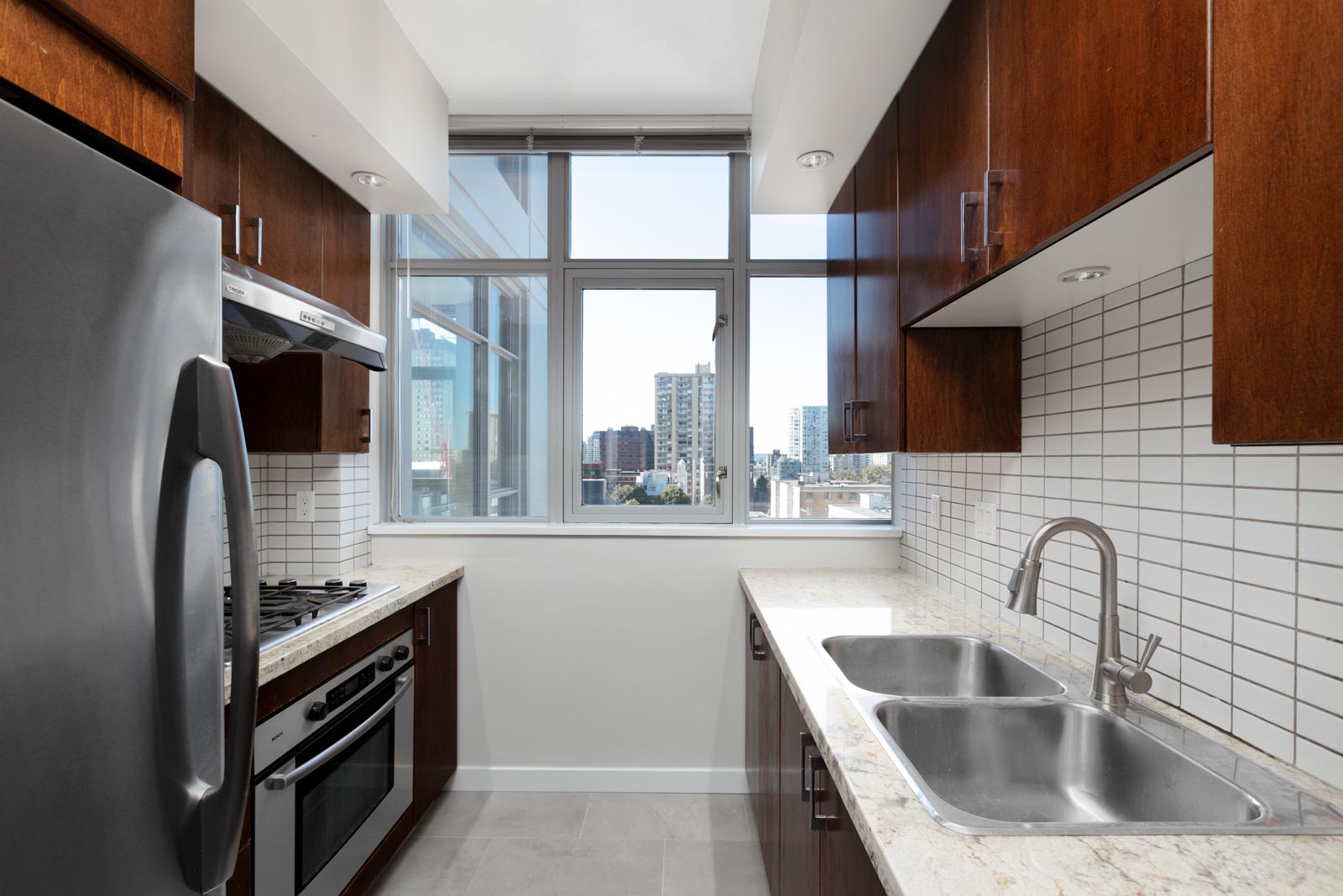 Modern kitchen with stainless steel appliances, dark wood cabinets, white tile backsplash, double sink, and a large window overlooking city buildings.