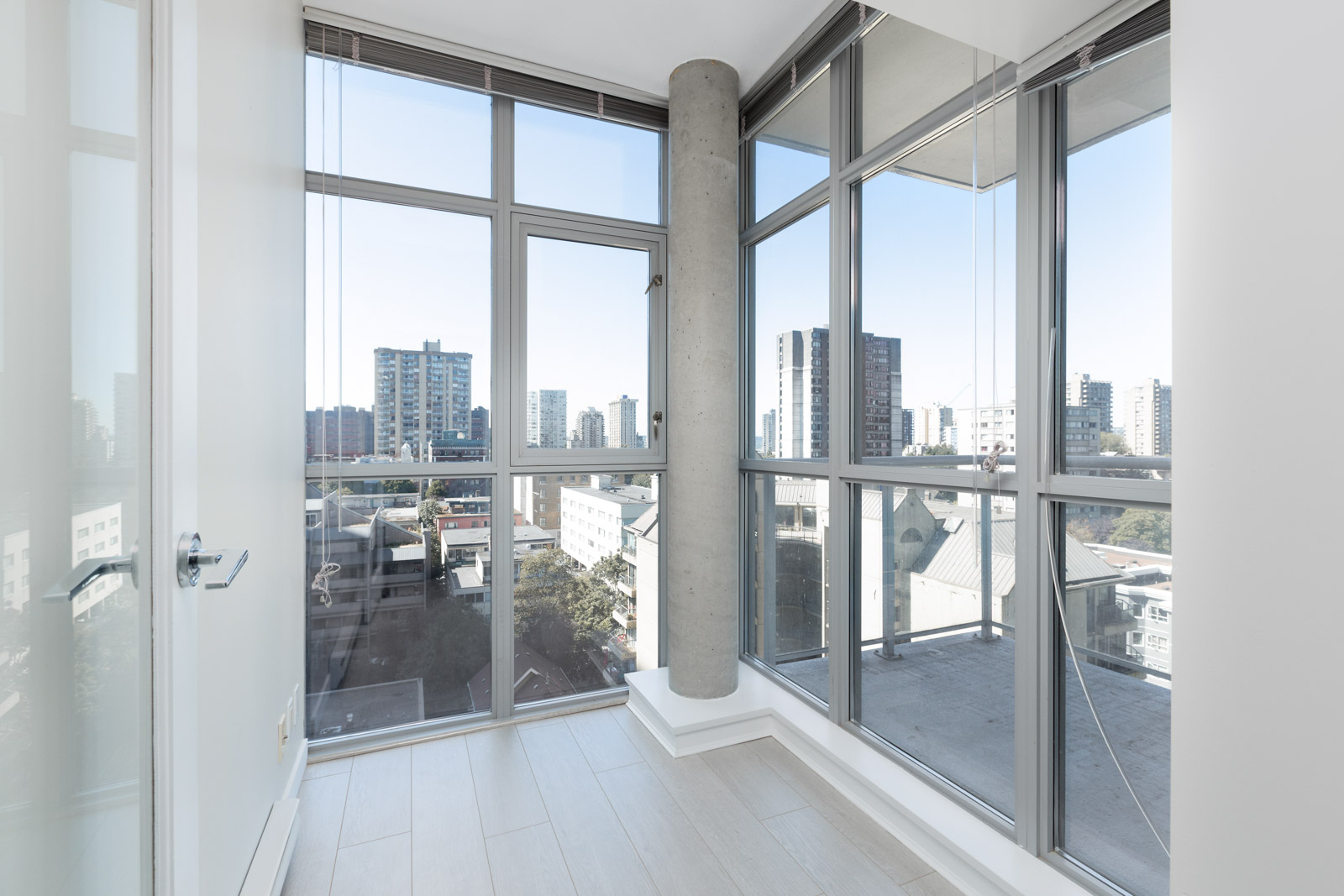 A small, empty corner room with floor-to-ceiling windows, a concrete pillar, light wood floors, and a cityscape view outside.