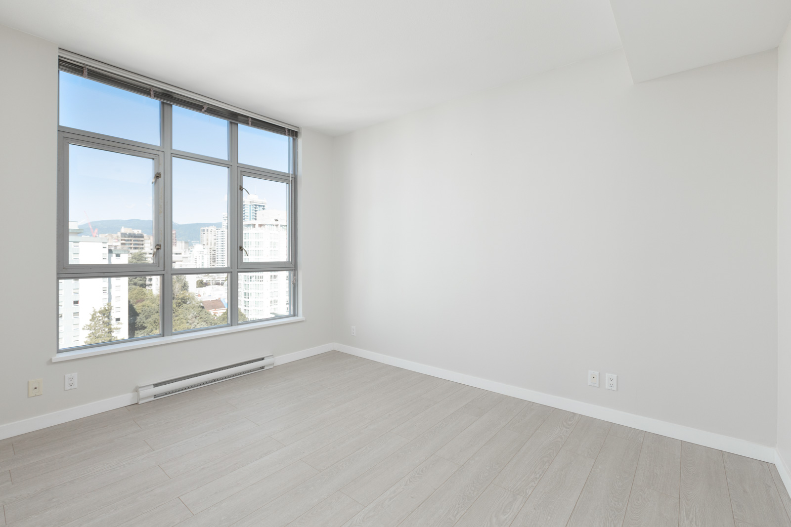 Empty room with light-colored wood flooring, white walls, and a large window offering a city view with buildings and mountains in the distance.