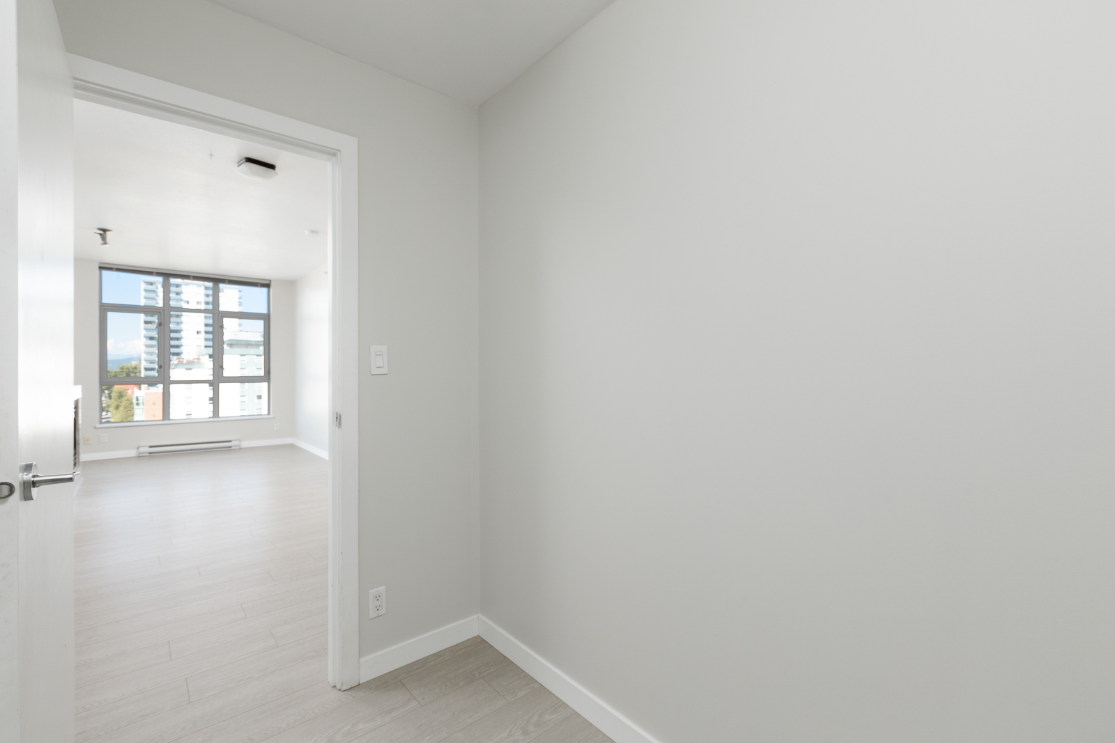 A view from a doorway into a bright, empty room with large windows, white walls, and light wood flooring in a modern apartment.