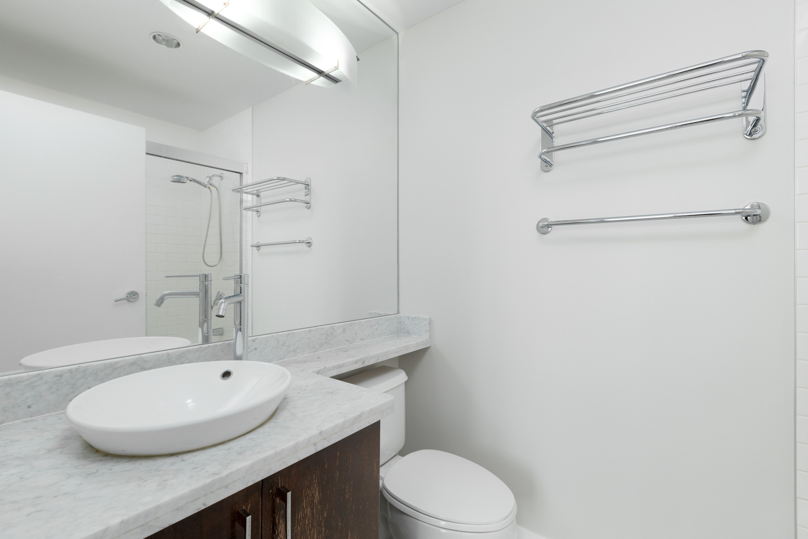 Modern bathroom with a round vessel sink, marble countertop, wall-mounted fixtures, toilet, towel bars, and a shower with glass door reflected in a large mirror.