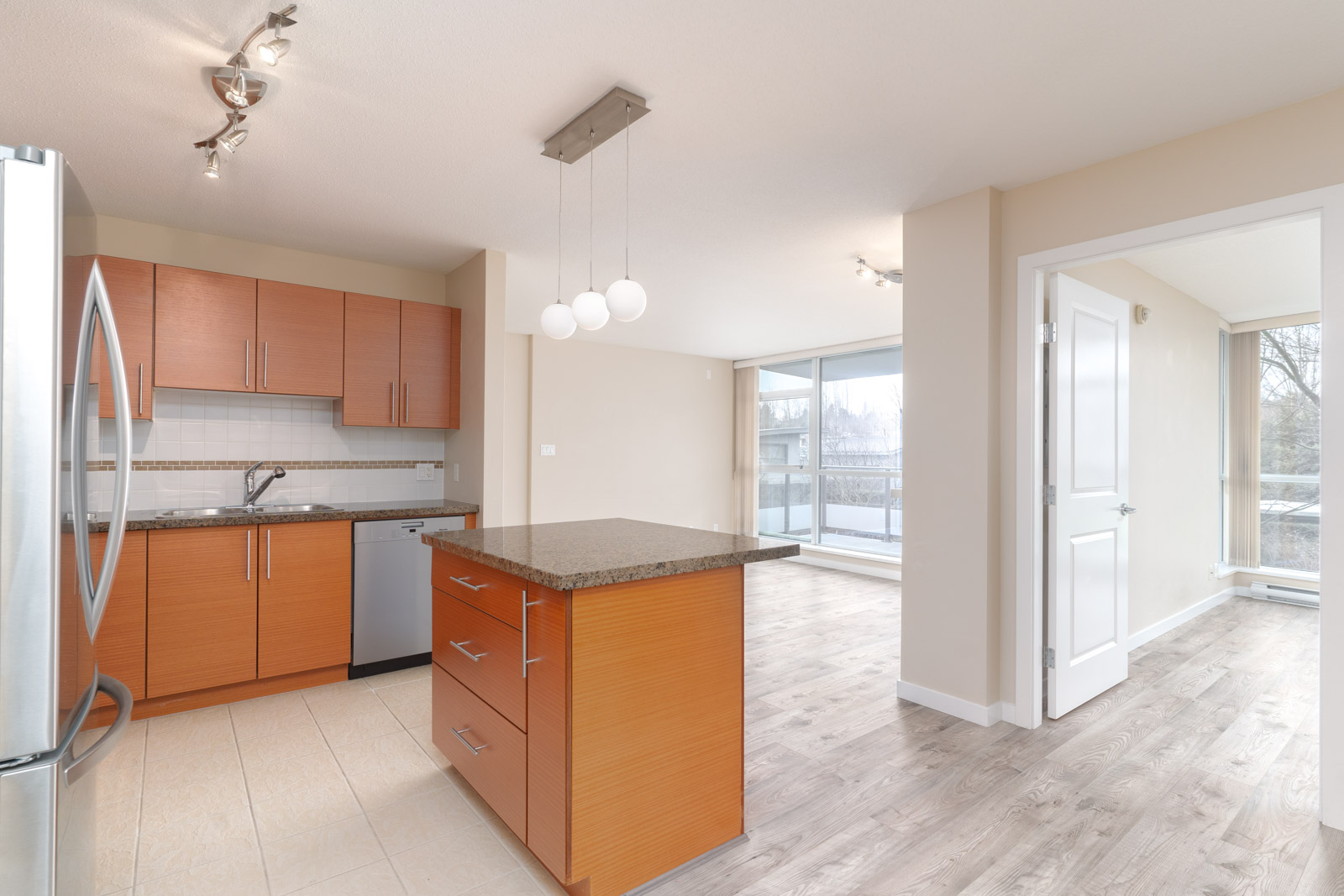 Modern kitchen with wood cabinets, granite island, stainless steel appliances, and open layout leading to a bright living area with large windows.