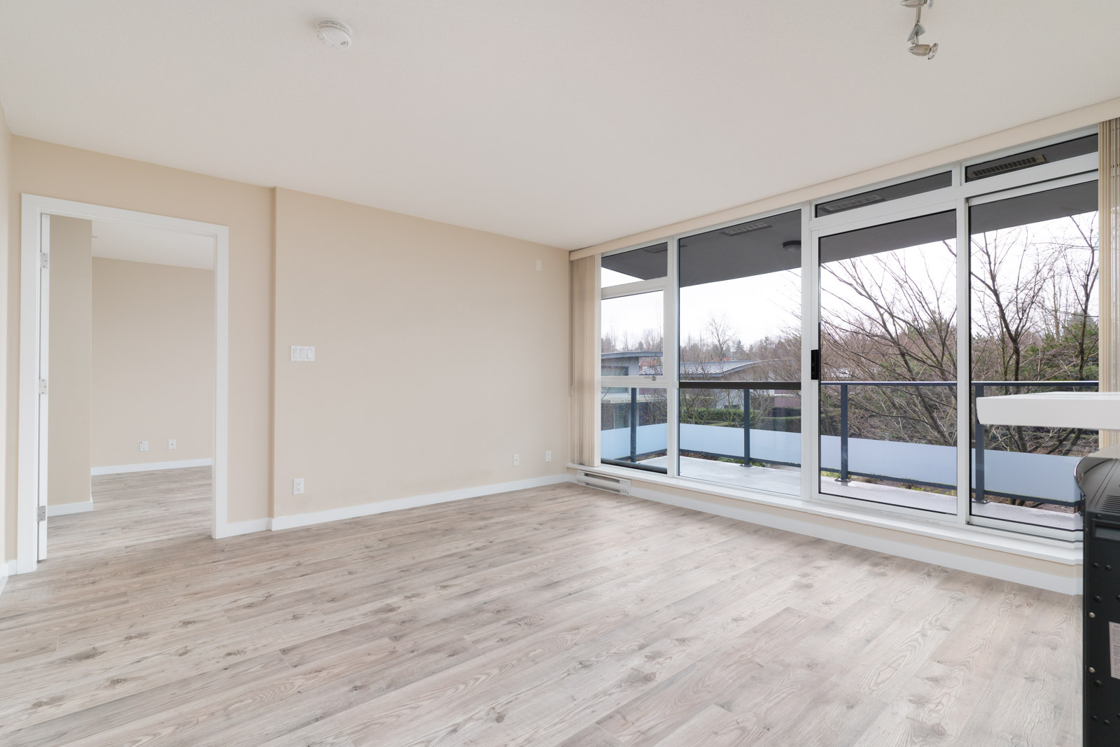 Unfurnished room with light wood floors, beige walls, floor-to-ceiling windows, and a sliding glass door leading to a balcony. Adjacent doorway opens into another room.