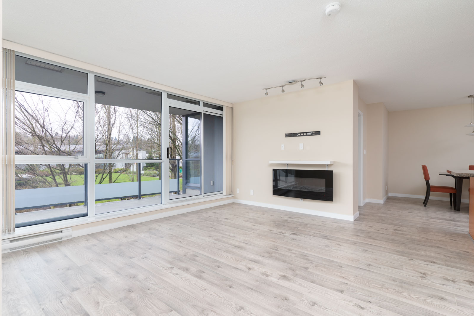 Unfurnished modern living room with large windows, light wood flooring, a wall-mounted fireplace, and an open view of a balcony with trees outside.