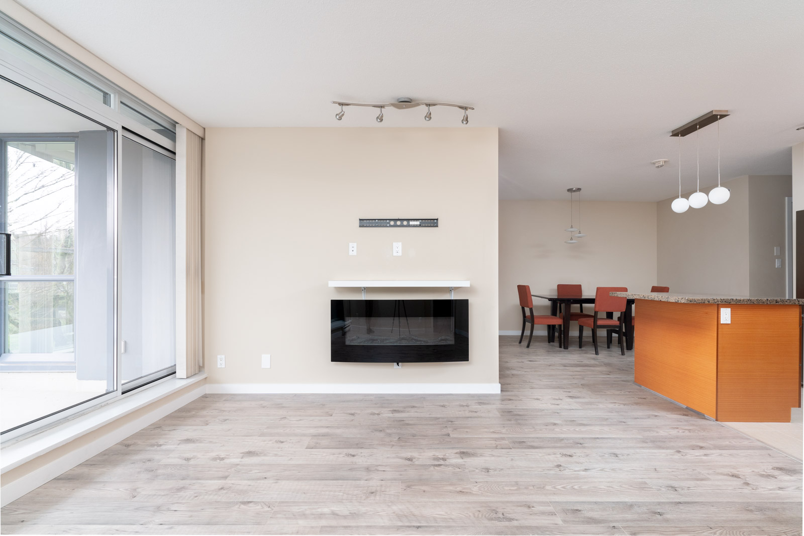 Modern, unfurnished living and dining area with large windows, light wood flooring, wall-mounted fireplace, and a dining table with red chairs near an orange kitchen island.