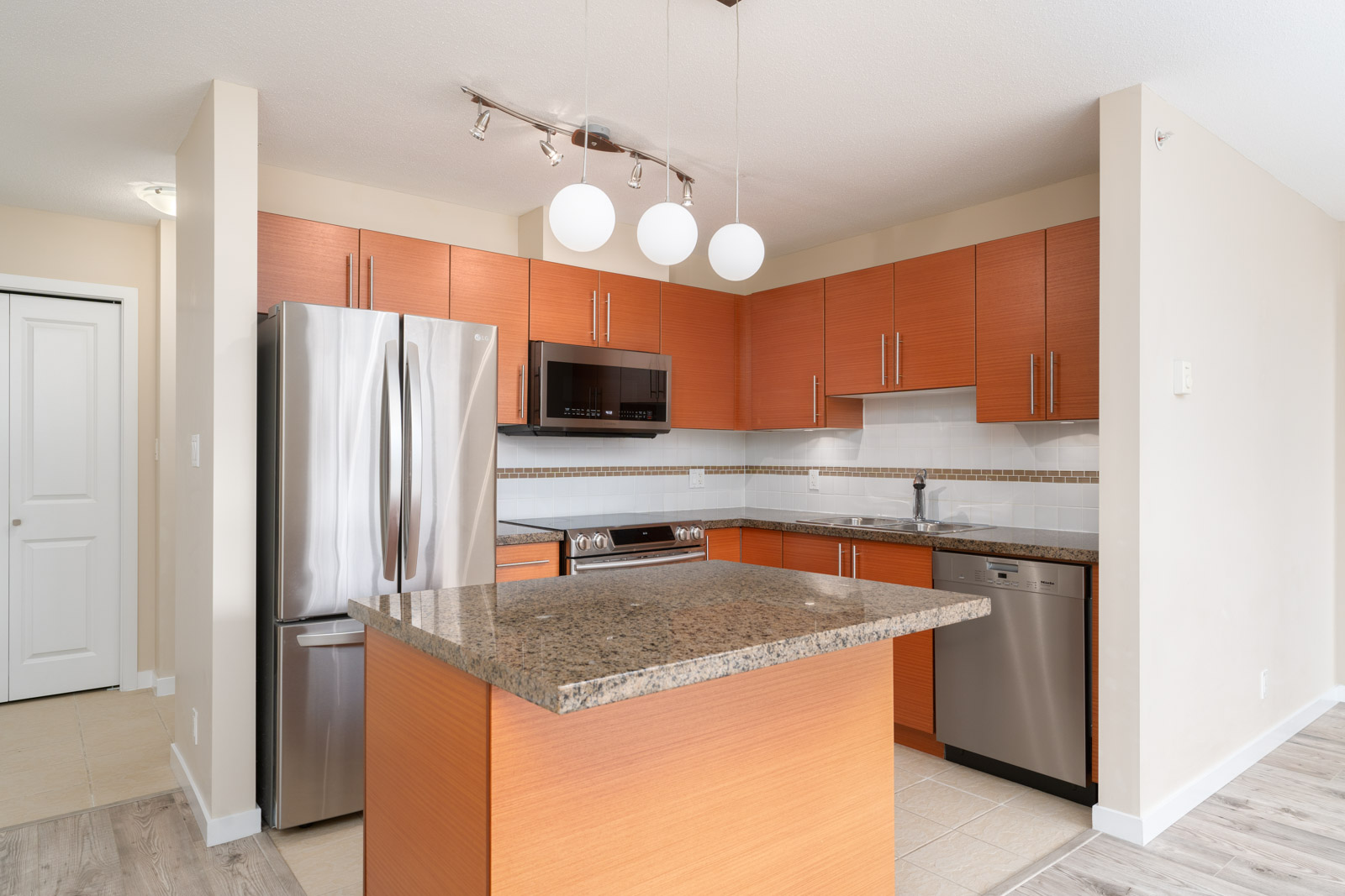 Modern kitchen with wood cabinets, stainless steel appliances, granite countertops, a center island, and pendant lights. Neutral walls and tile flooring are also visible.