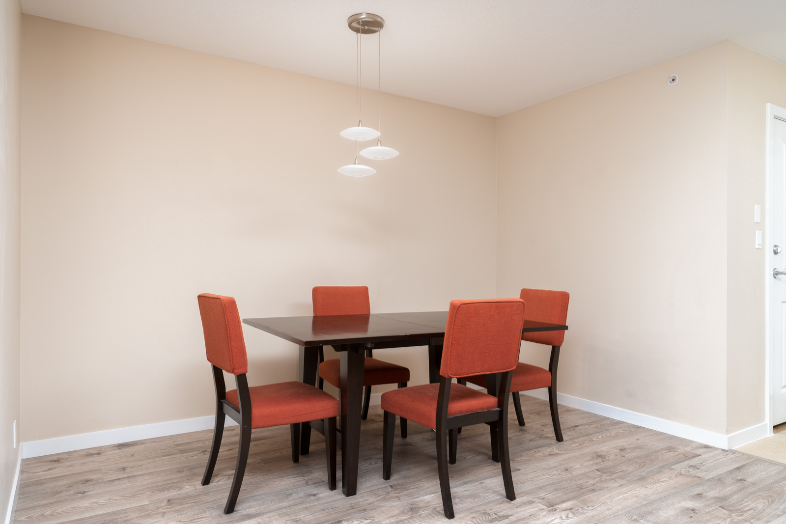 Dining area with a dark wooden table, four orange upholstered chairs, wood-style flooring, beige walls, and a modern ceiling light fixture.