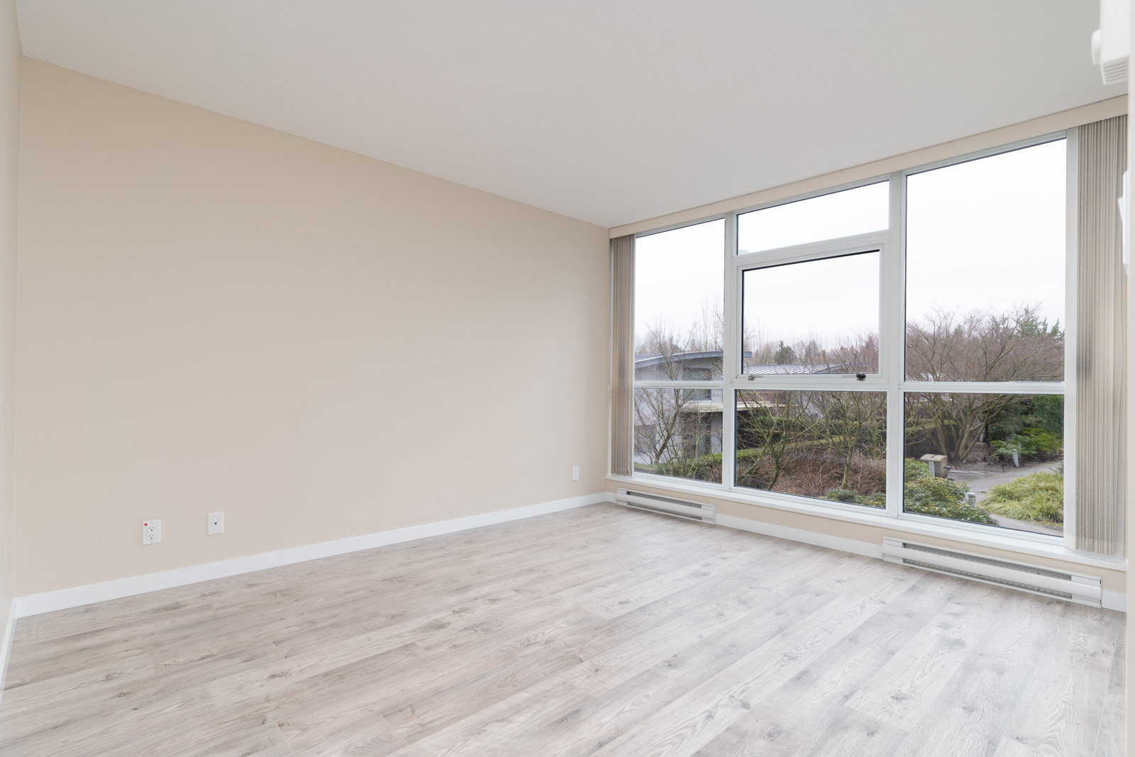 Empty room with light-colored walls, large floor-to-ceiling windows, and light wood flooring, overlooking a garden and trees outside.
