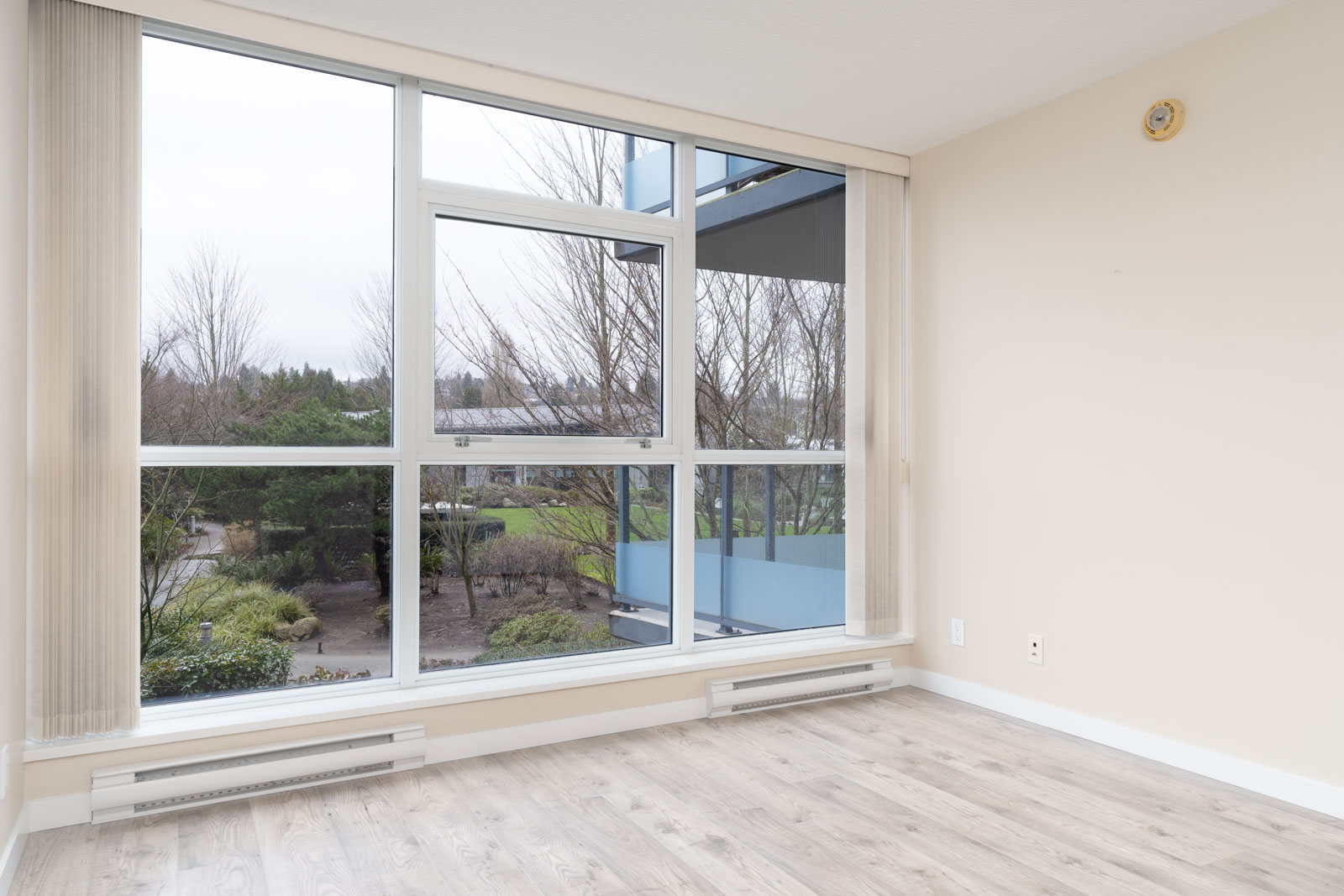 Empty room with light-colored walls and wood flooring, featuring large windows overlooking an outdoor garden with trees and shrubs.