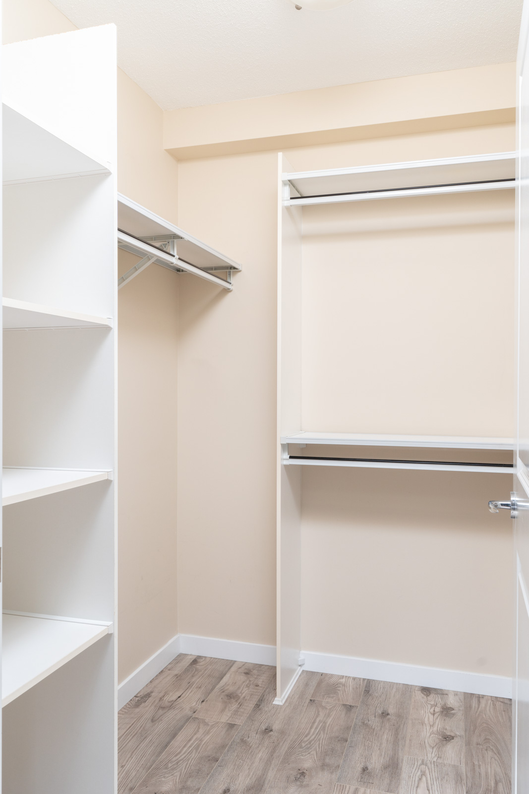 Empty walk-in closet with beige walls, white shelving units, hanging rods, and wood-patterned flooring.