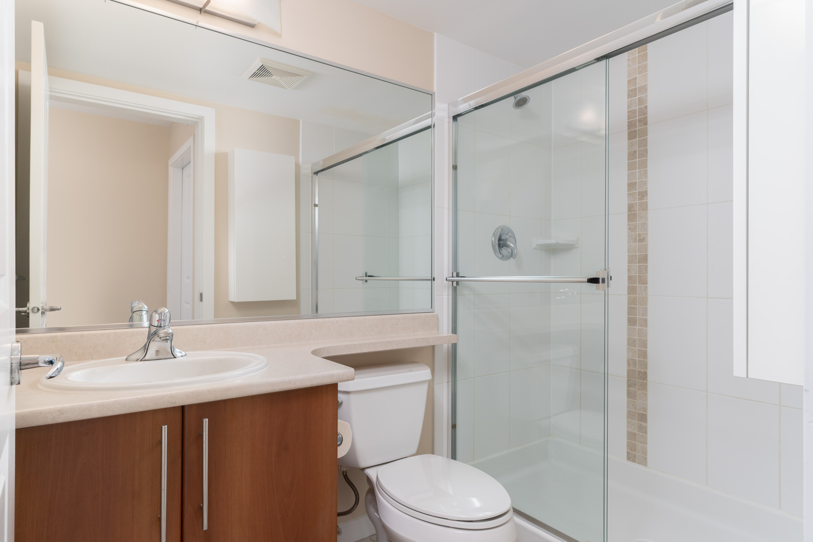 Modern bathroom with a sink, wooden vanity, toilet, and glass-enclosed shower with beige tiled accent. Large mirror above the sink and overhead lighting are visible.