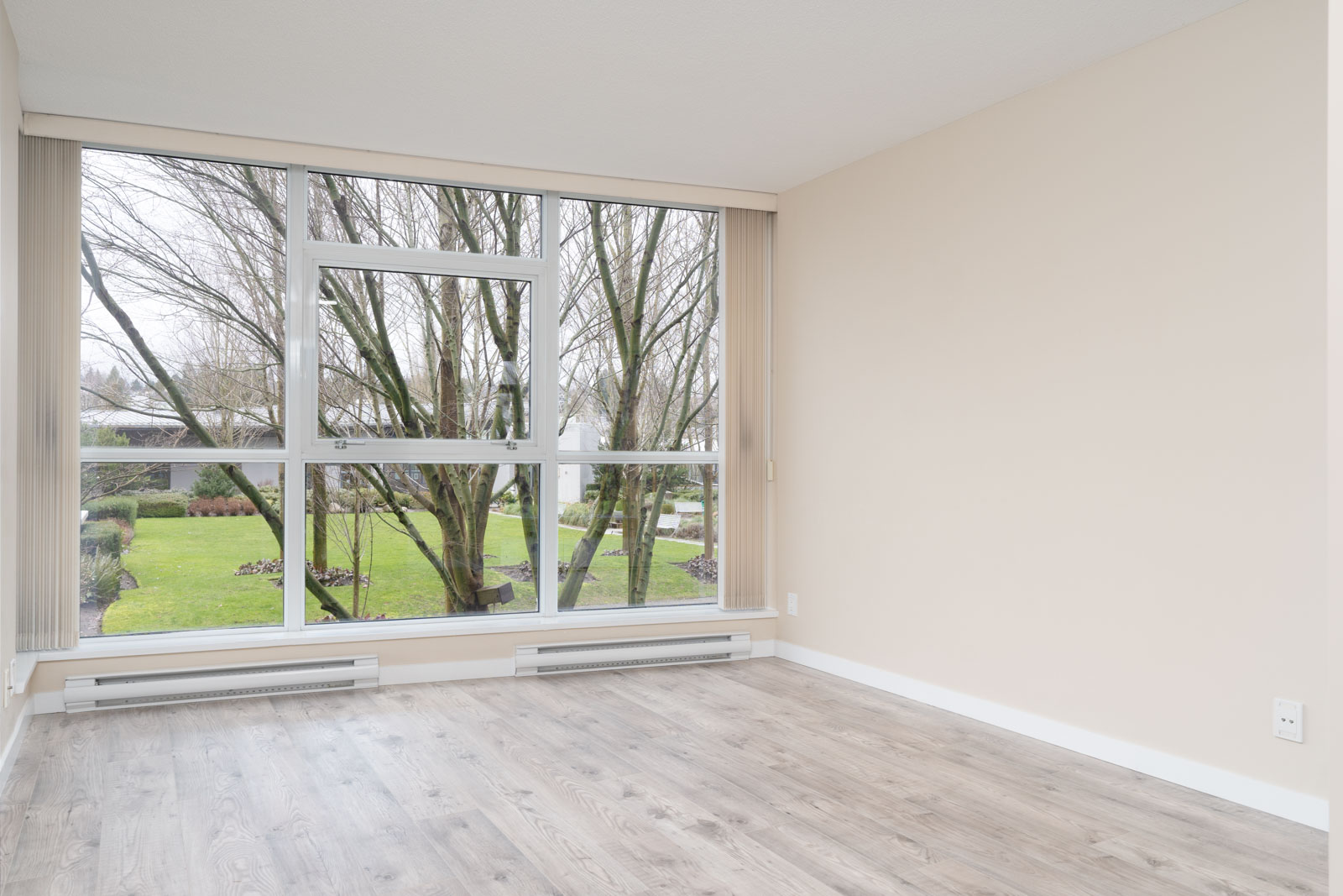 Unfurnished room with light wood flooring, beige walls, and large windows overlooking a grassy yard with trees.