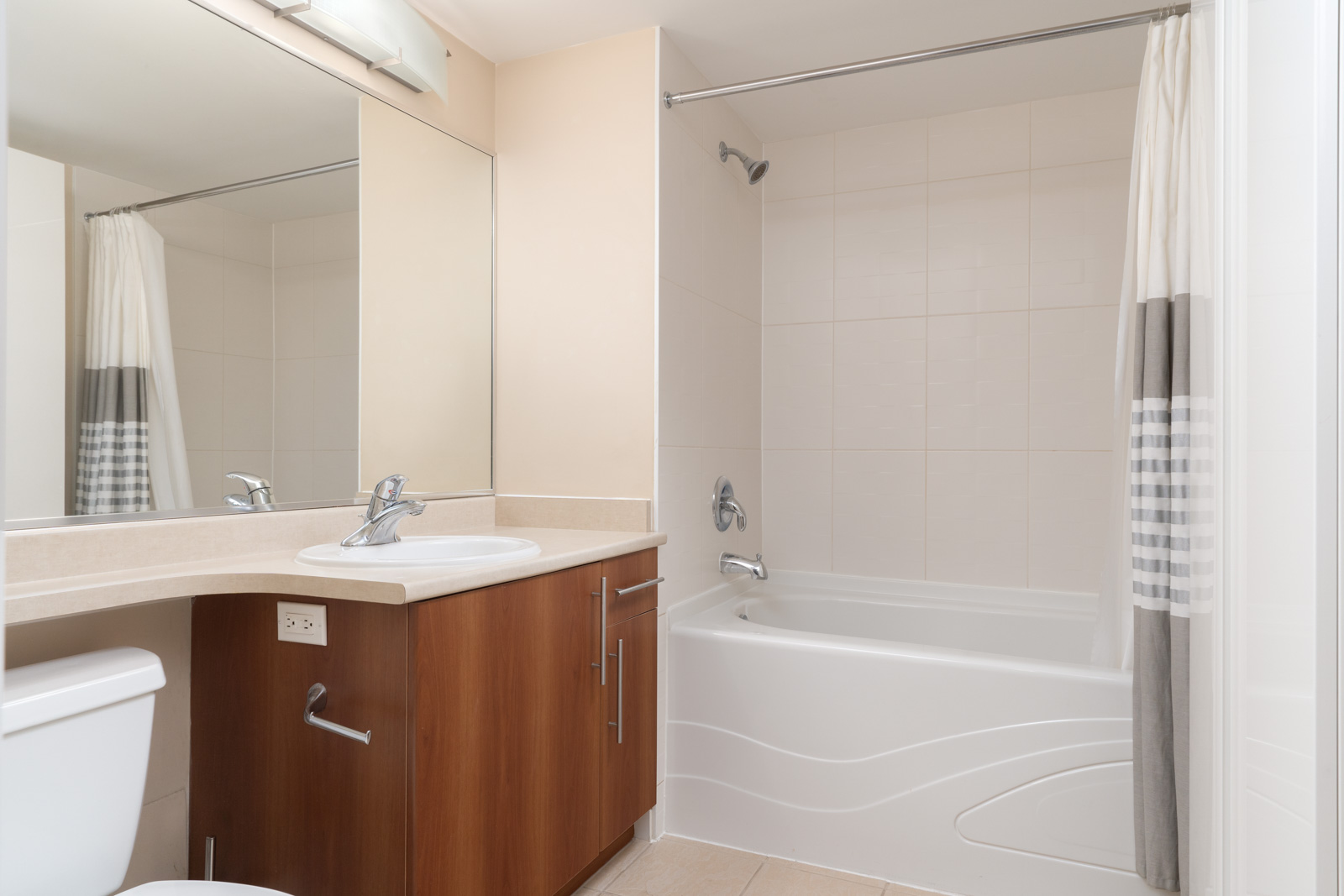 Modern bathroom with a white bathtub and shower, beige walls, large mirror, wooden vanity with sink, and striped shower curtain.