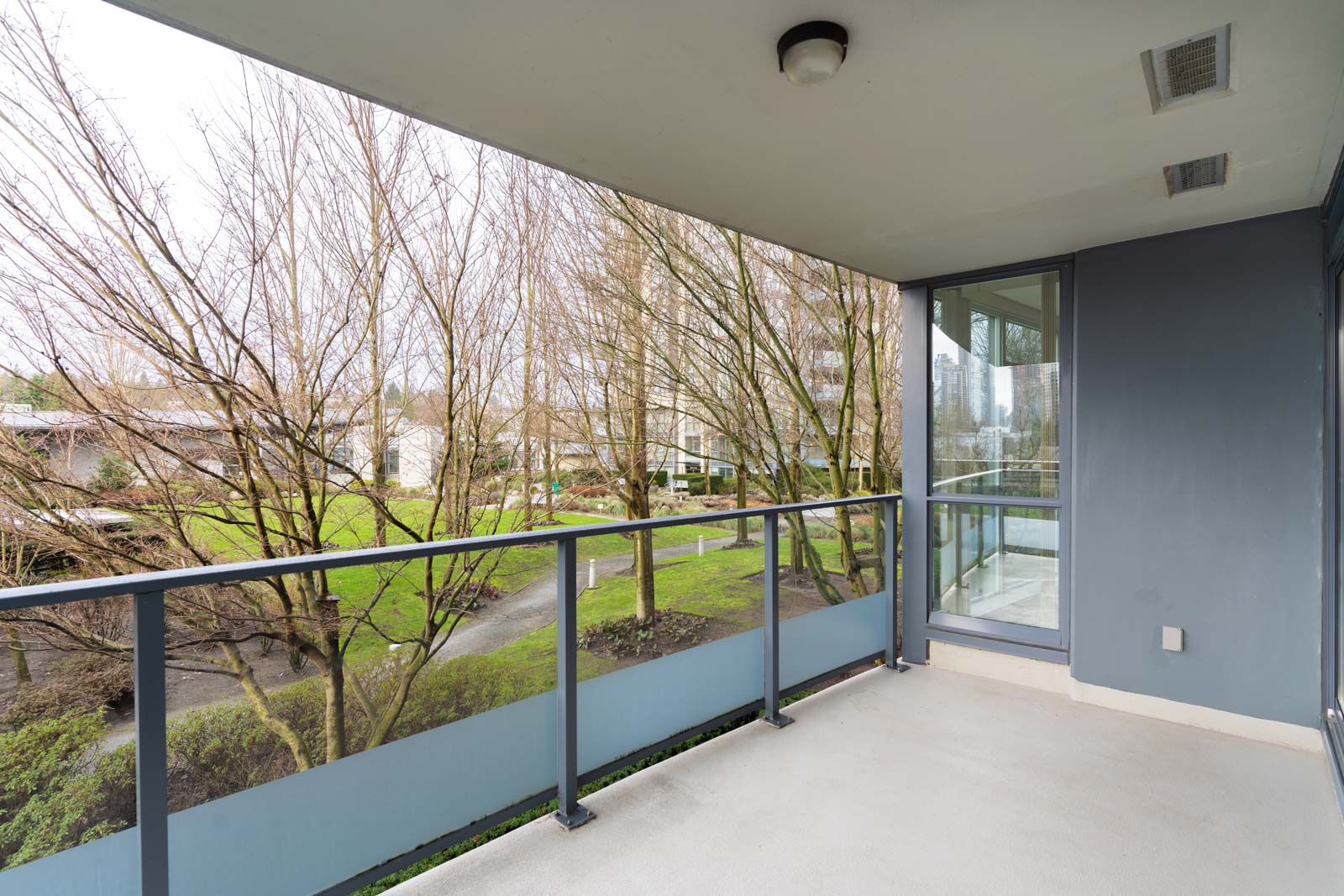 A covered balcony with glass railing overlooks a landscaped courtyard with leafless trees and walkways on an overcast day.