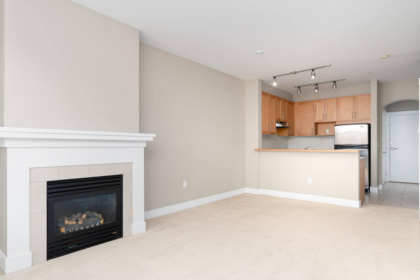Unfurnished apartment living area with beige walls, a gas fireplace, carpeted floor, and an open kitchen featuring wooden cabinets and stainless steel appliances.