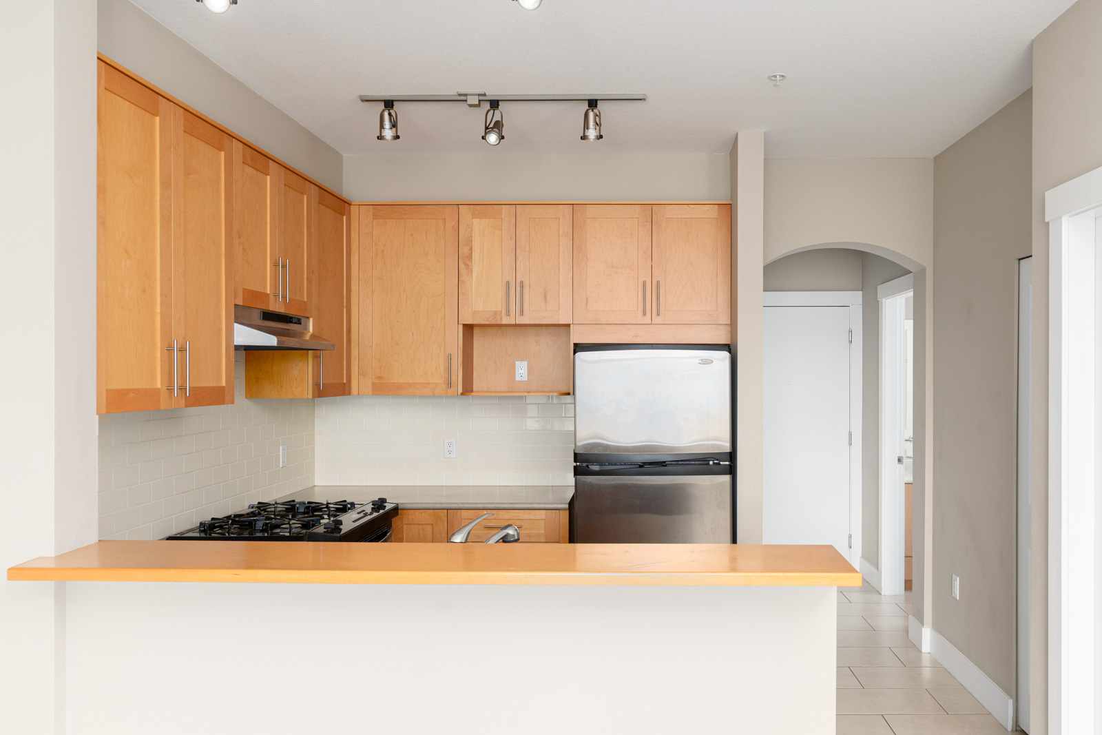 Modern kitchen with light wood cabinets, stainless steel appliances, gas stove, white tile backsplash, and beige walls, viewed from across a breakfast bar.