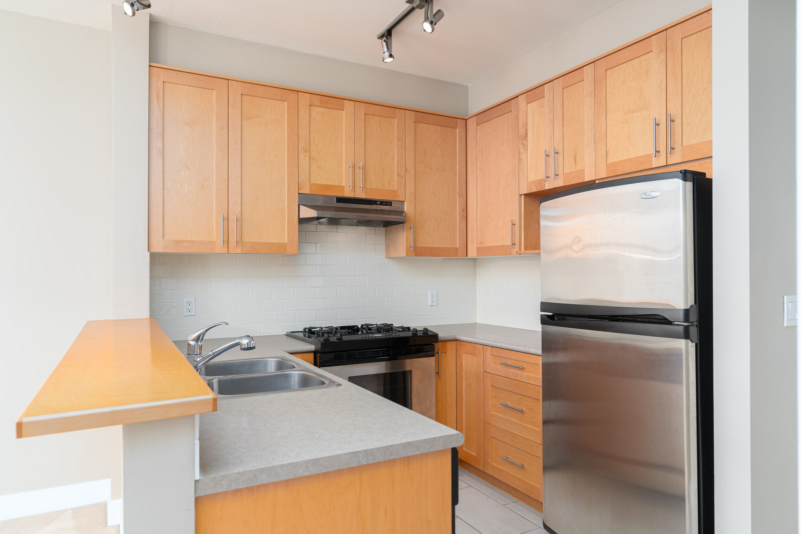 Modern kitchen with light wood cabinets, stainless steel refrigerator, gas stove, double sink, and light-colored tile backsplash.