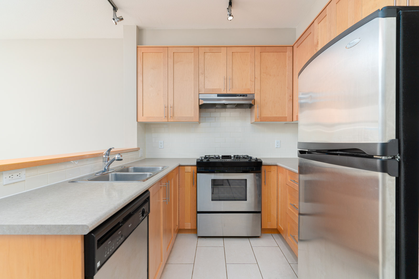 Modern kitchen with light wood cabinets, stainless steel refrigerator, oven, dishwasher, gas stove, and double sink on a gray countertop.