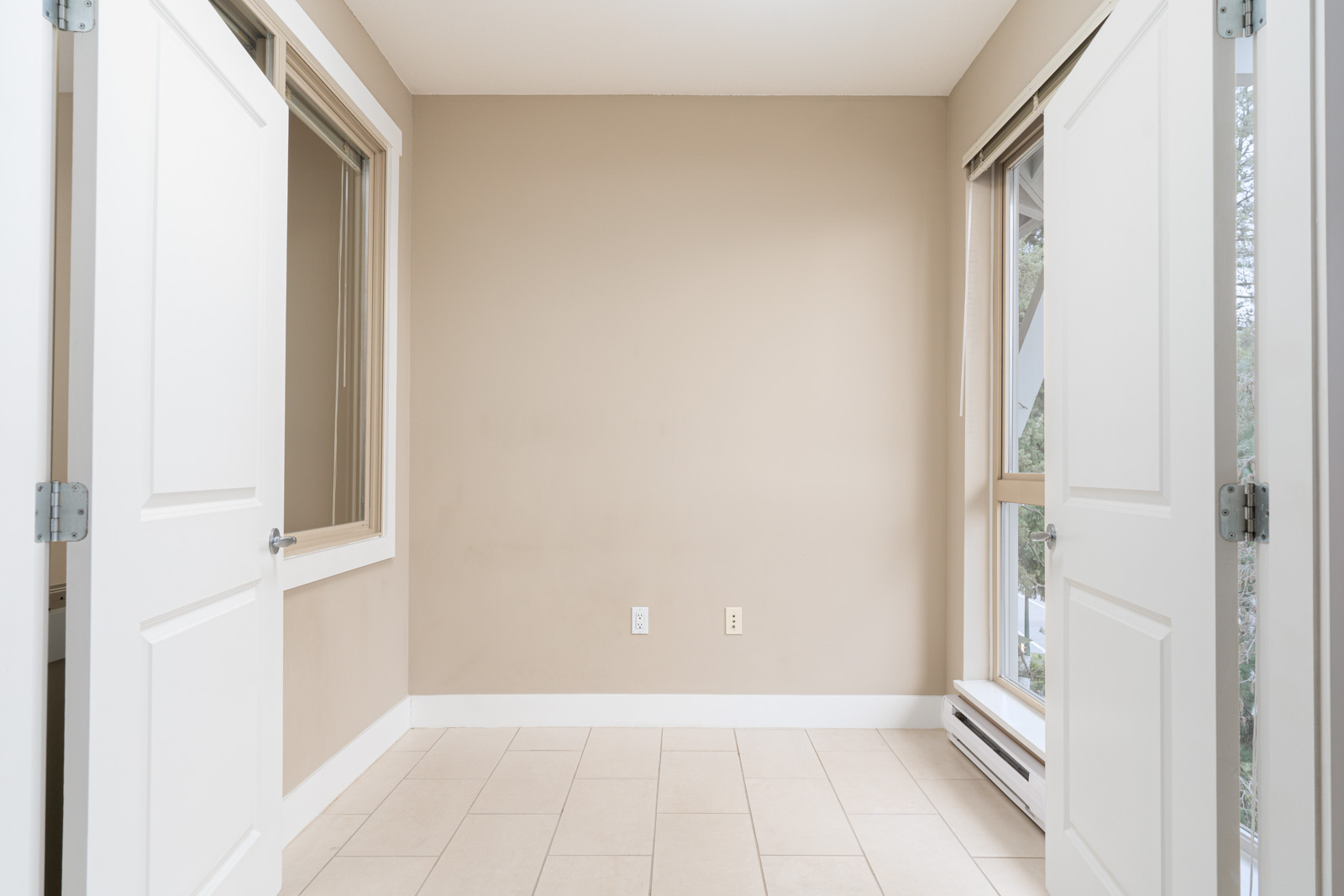 Empty room with beige walls, tiled floor, two open white doors, and two windows letting in natural light.