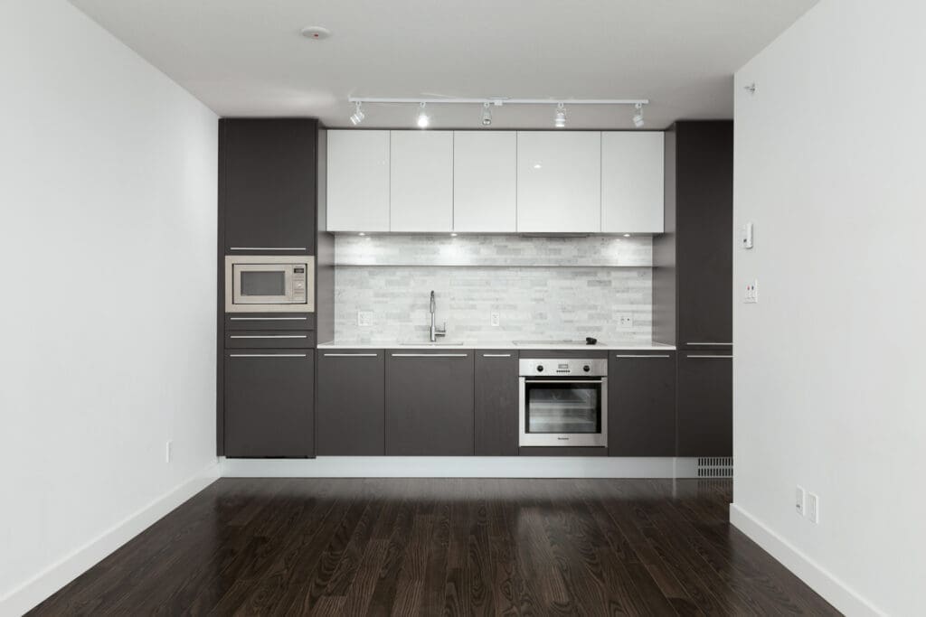 Modern kitchen with dark wood floors, grey and white cabinets, built-in microwave, oven, sink, and a tiled backsplash under track lighting.