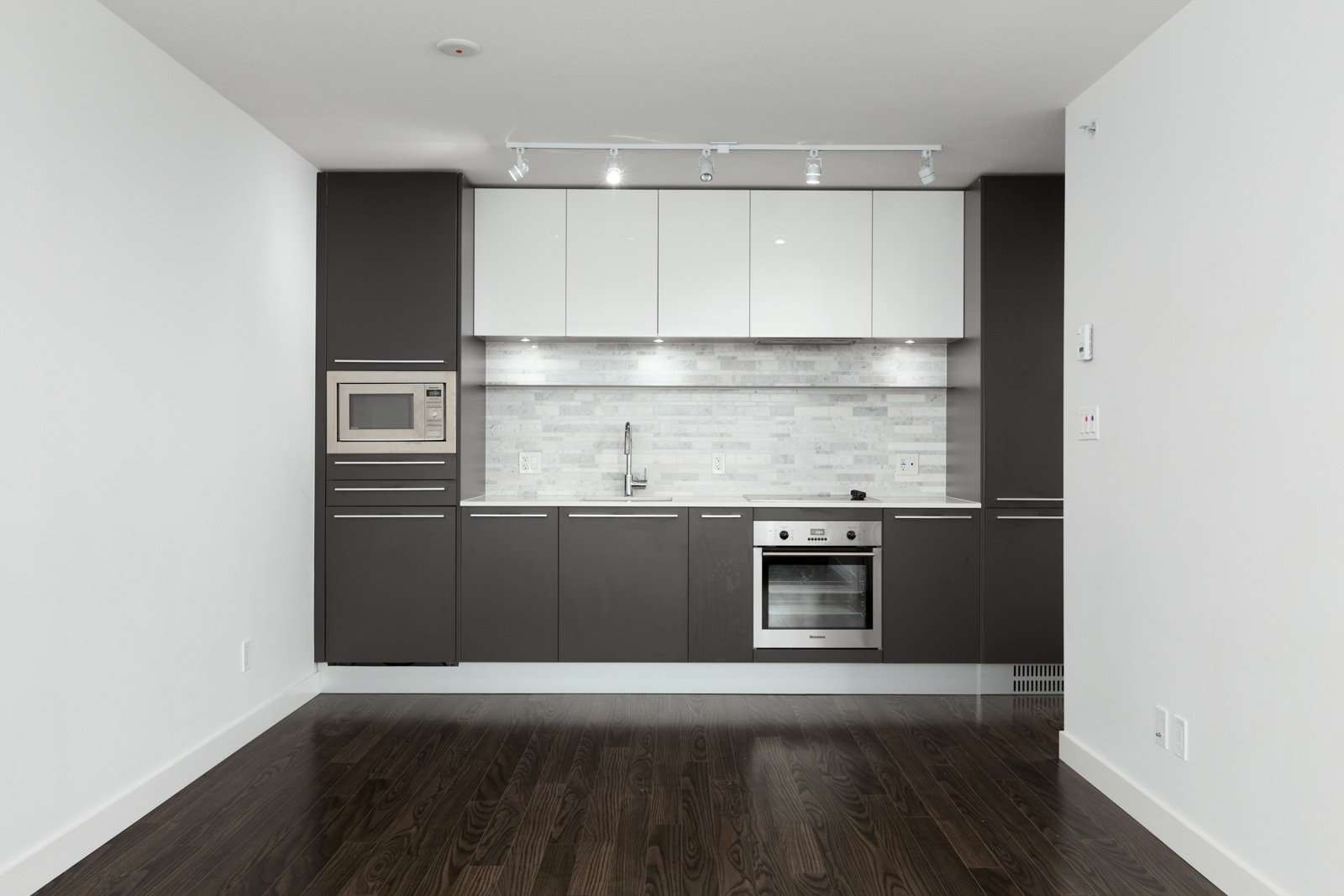 Modern kitchen with dark wood floors, grey and white cabinets, built-in microwave, oven, sink, and a tiled backsplash under track lighting.