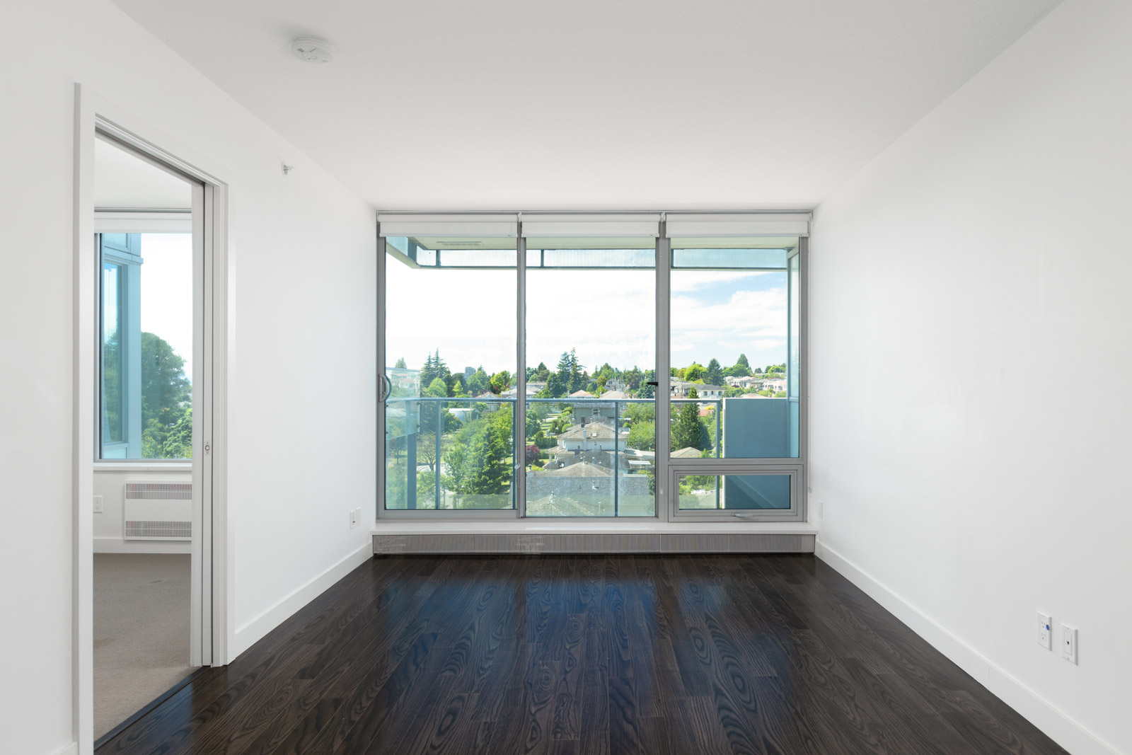 Unfurnished apartment room with dark wood floors, large floor-to-ceiling windows, a glass balcony door, and a view of trees and buildings outside. Adjacent room visible on the left.