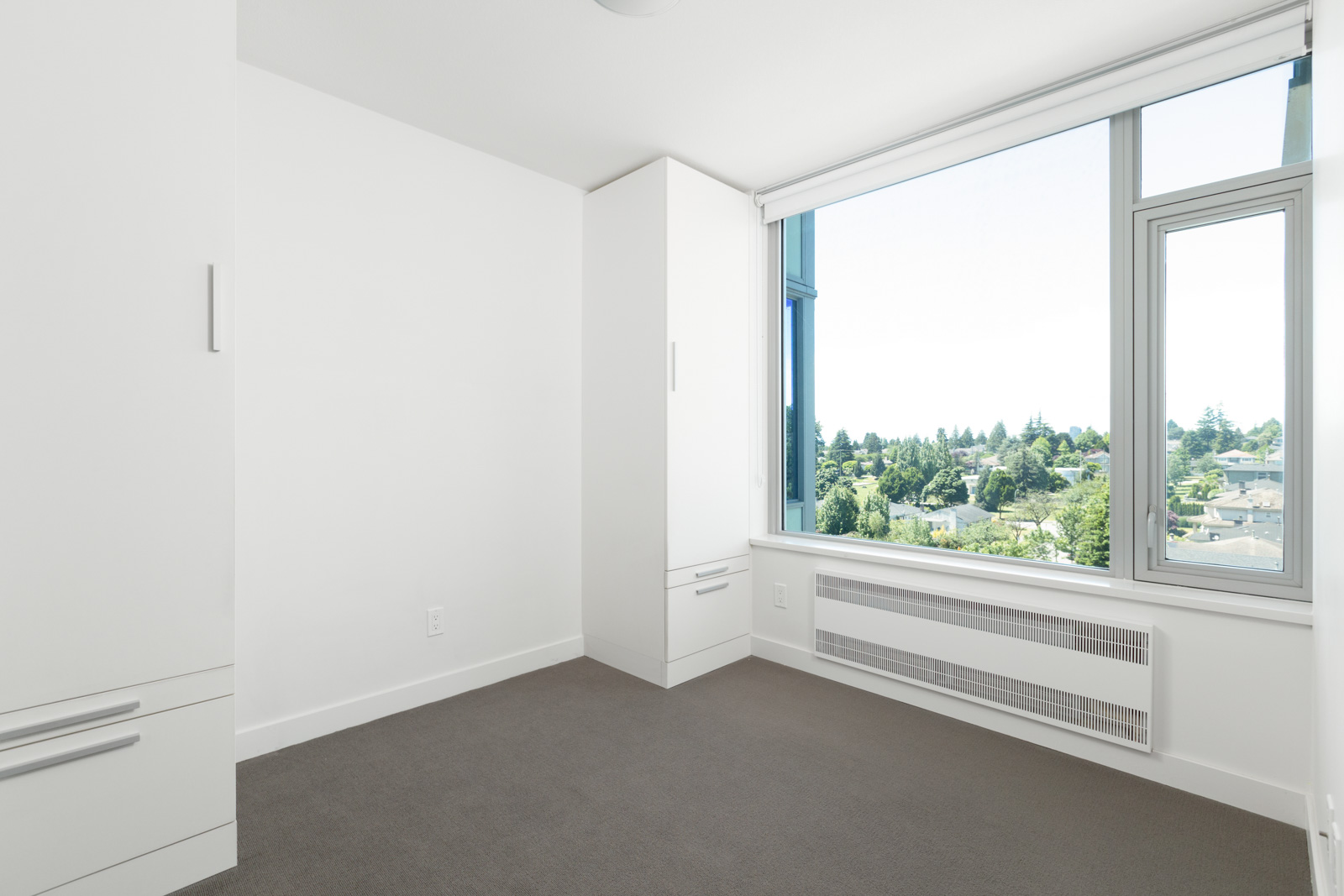 Empty, unfurnished room with white walls, built-in cabinets, and a large window offering a view of trees and buildings outside.