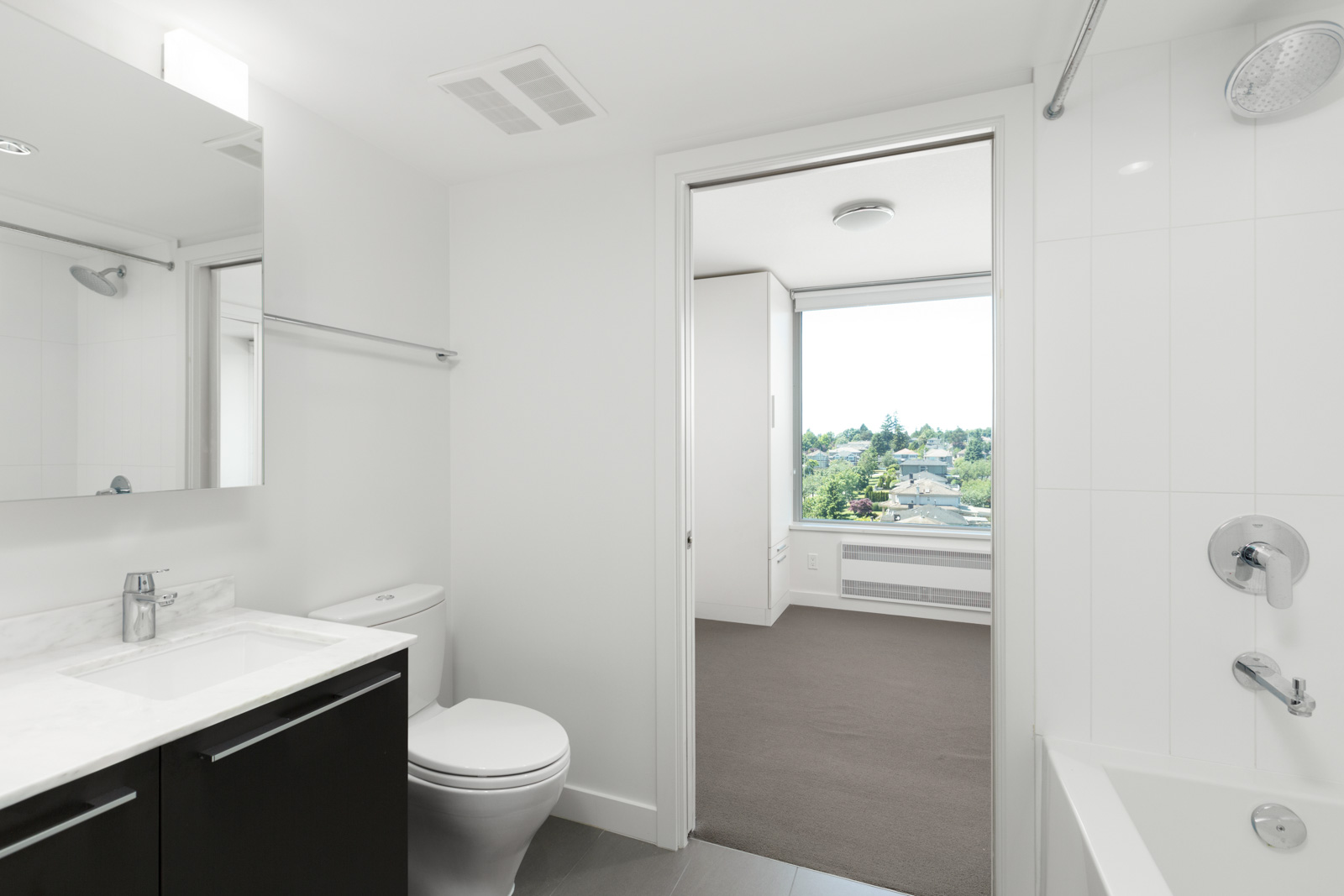 Modern white bathroom with a sink, toilet, and bathtub, leading into a carpeted bedroom with a large window and a view of trees and buildings outside.