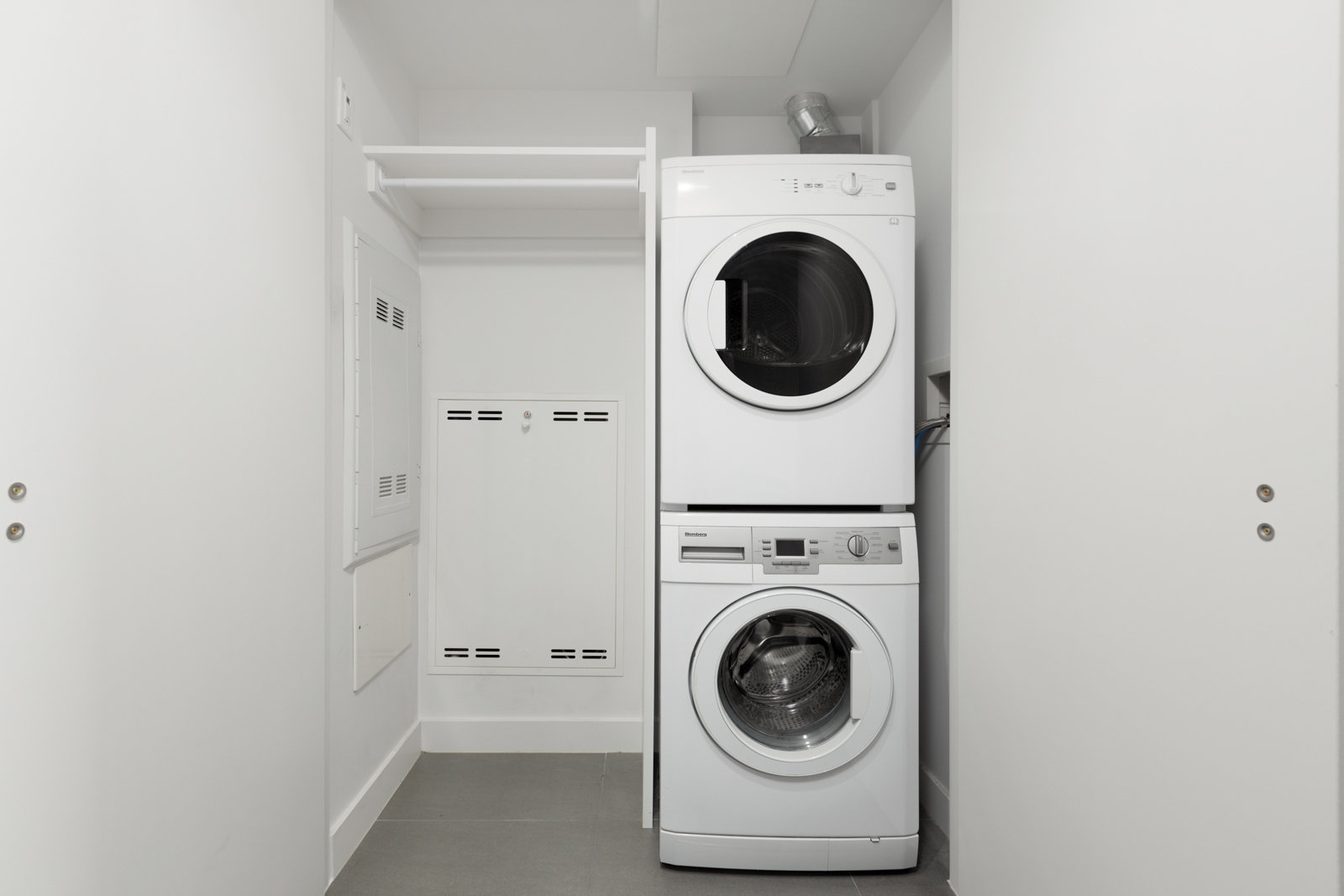 A stacked white washer and dryer set in a small, white laundry closet with open shelves and gray tile flooring.