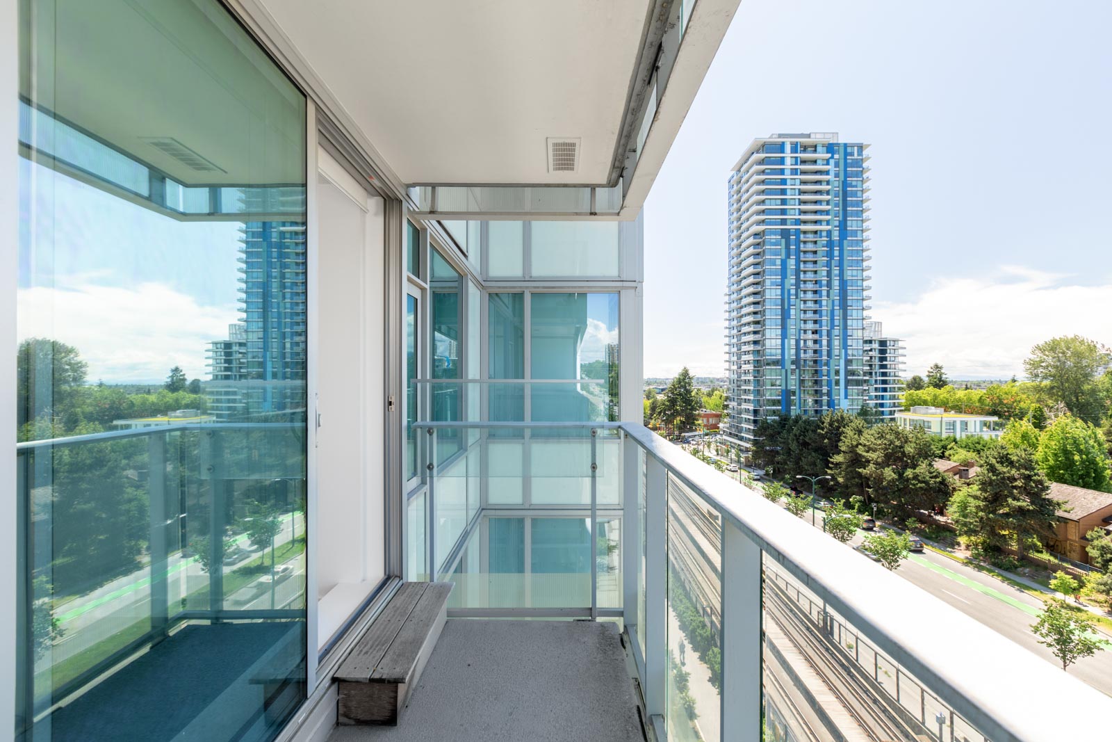 A modern apartment balcony with glass railings overlooks a street, trees, and several tall residential buildings under a clear sky.