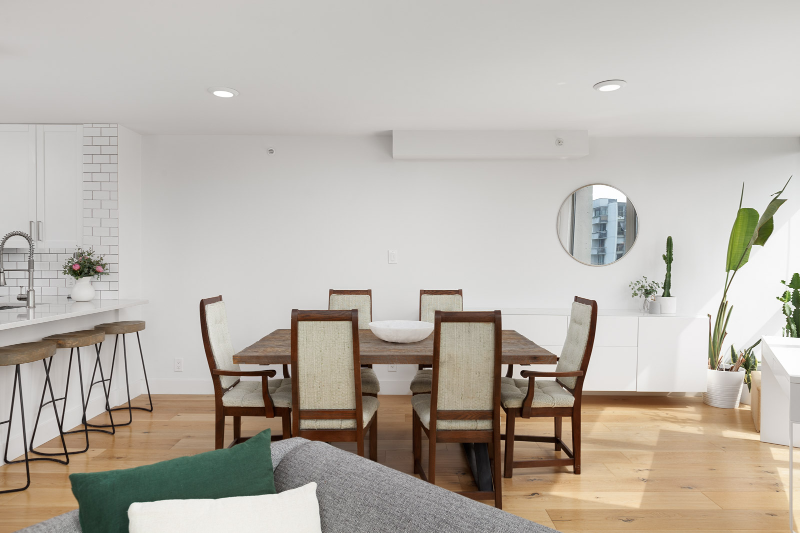 Modern dining area with a wooden table, six chairs, a round mirror on the wall, and plants near a large window in a bright, minimalist apartment.