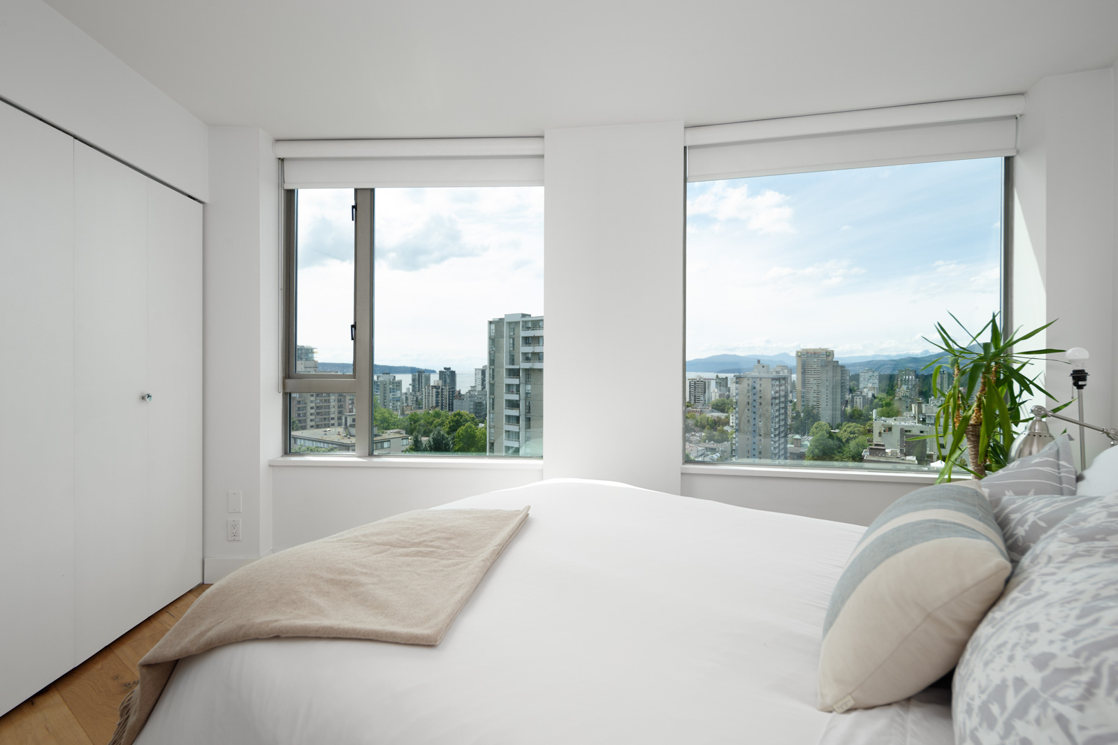 Bright bedroom with large windows, a neatly made bed, and a cityscape view featuring tall buildings and distant mountains.
